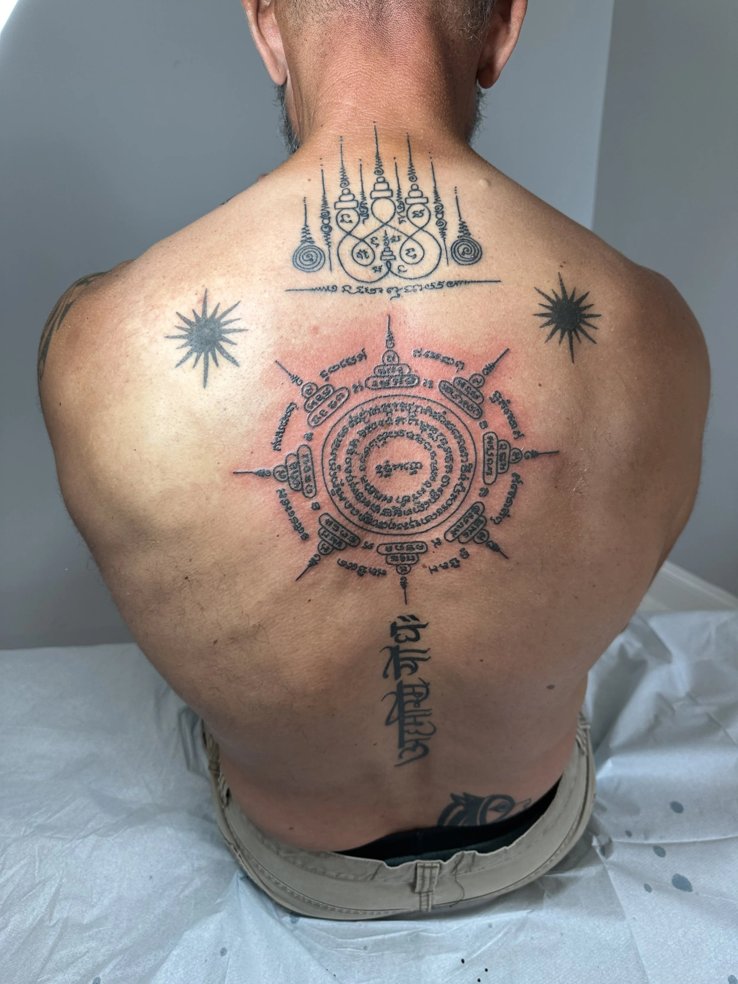 Tattoo on the back of a person with a large, intricate, circular pattern with script and symbols, flanked by two star-like designs.