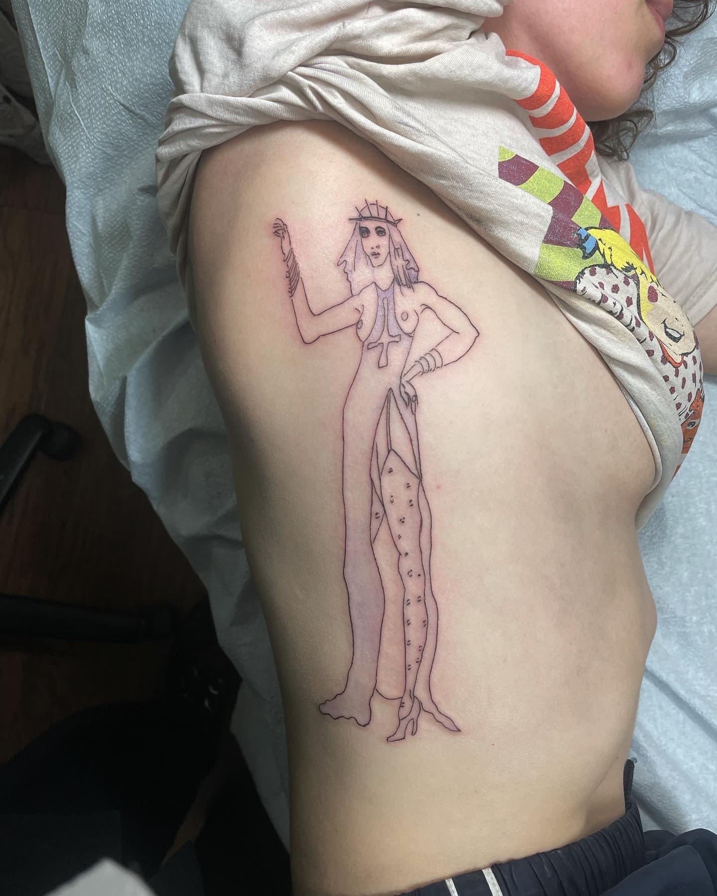 Temporary tattoo of a stylized female figure with a halo and spiked crown, wearing a gown with a high slit, standing on a leg made of a plant or tree trunk.