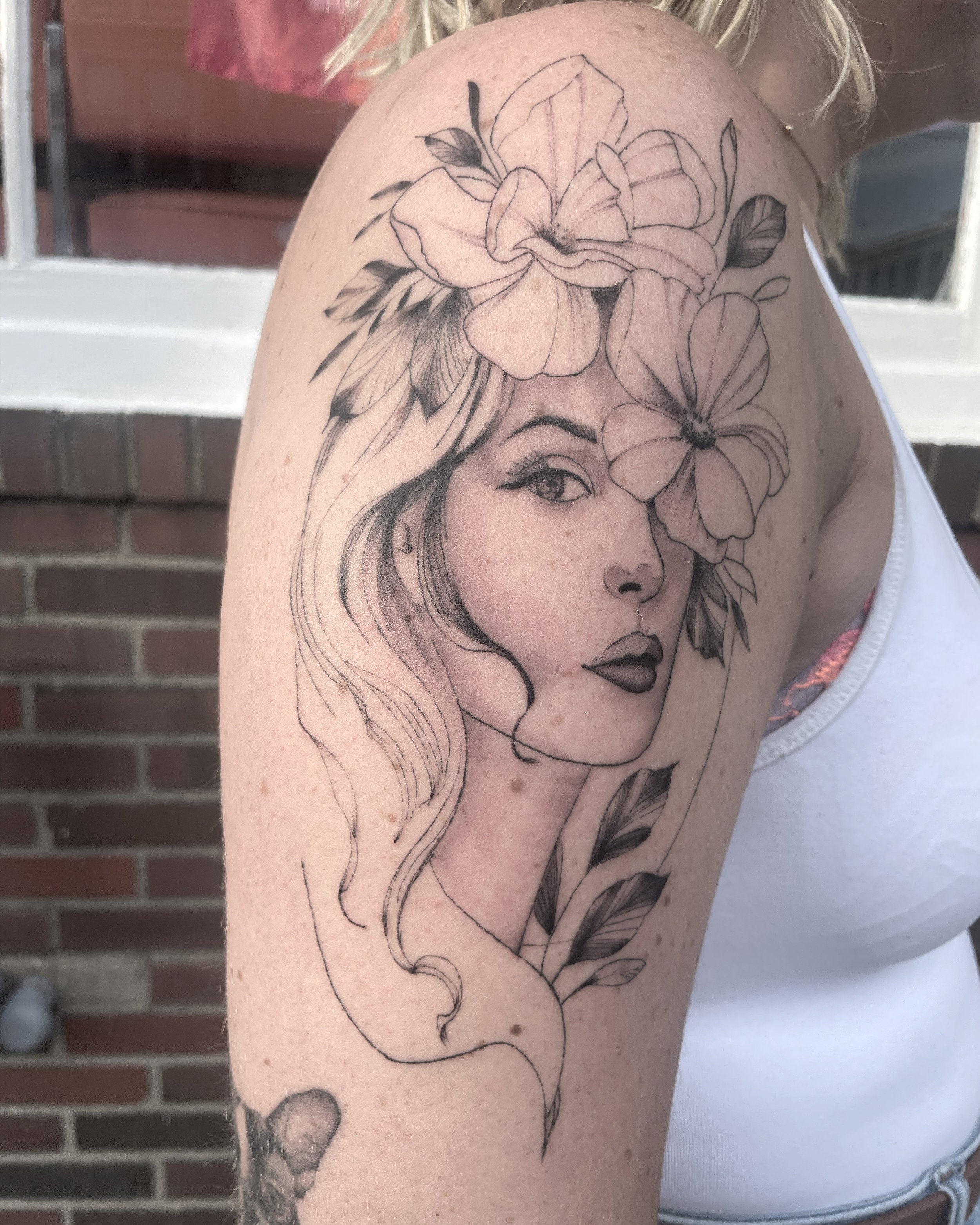 Tattoo of a woman's face with flowers and leaves on her head on person's upper arm.