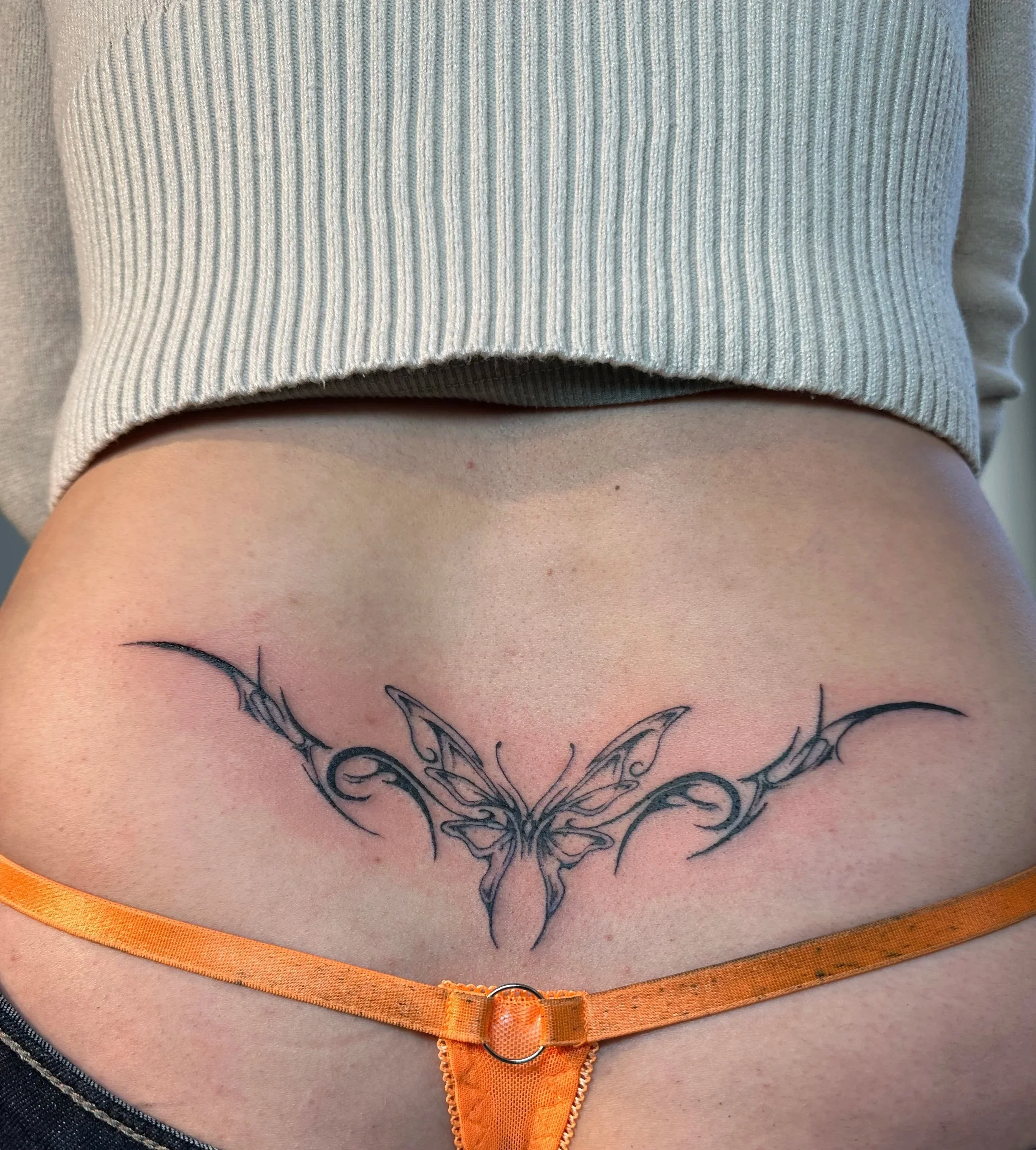 A lower back tattoo of a butterfly with outstretched wings, featuring intricate line work and tribal design elements, beneath a gray ribbed sweater and above orange underwear.