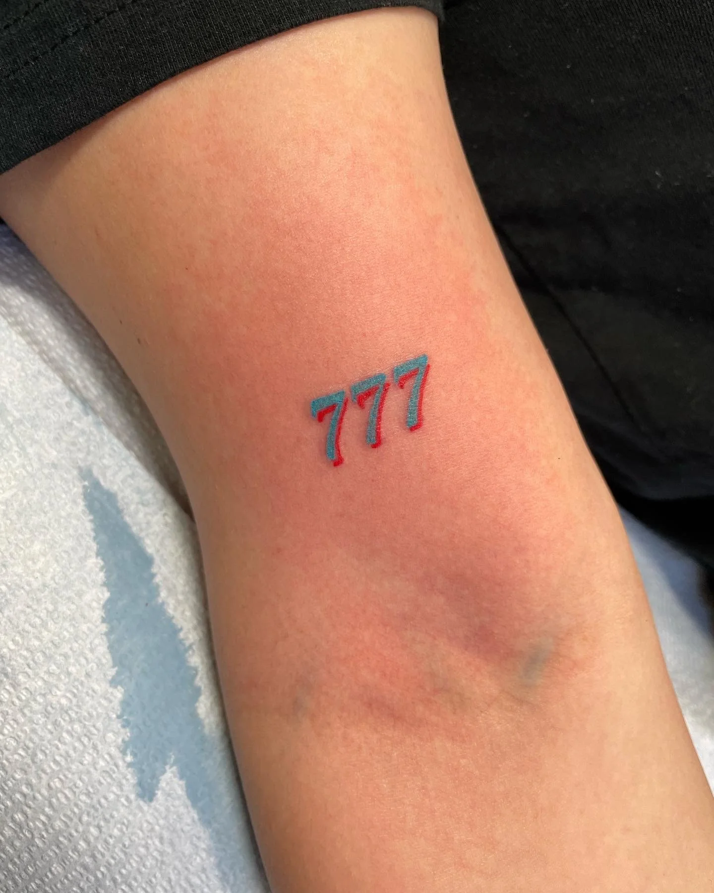 A tattoo of the number 777 in blue with red shadows on a person's arm.