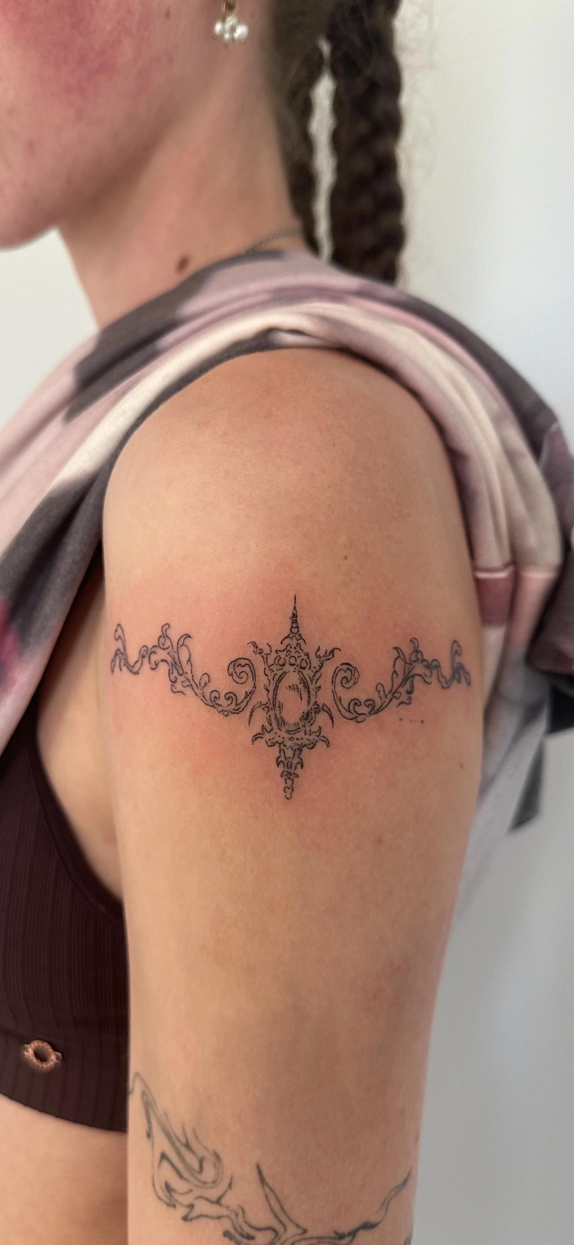 Close-up of a woman's upper arm and shoulder showing a black line tattoo with intricate, ornate patterns and a central oval design.