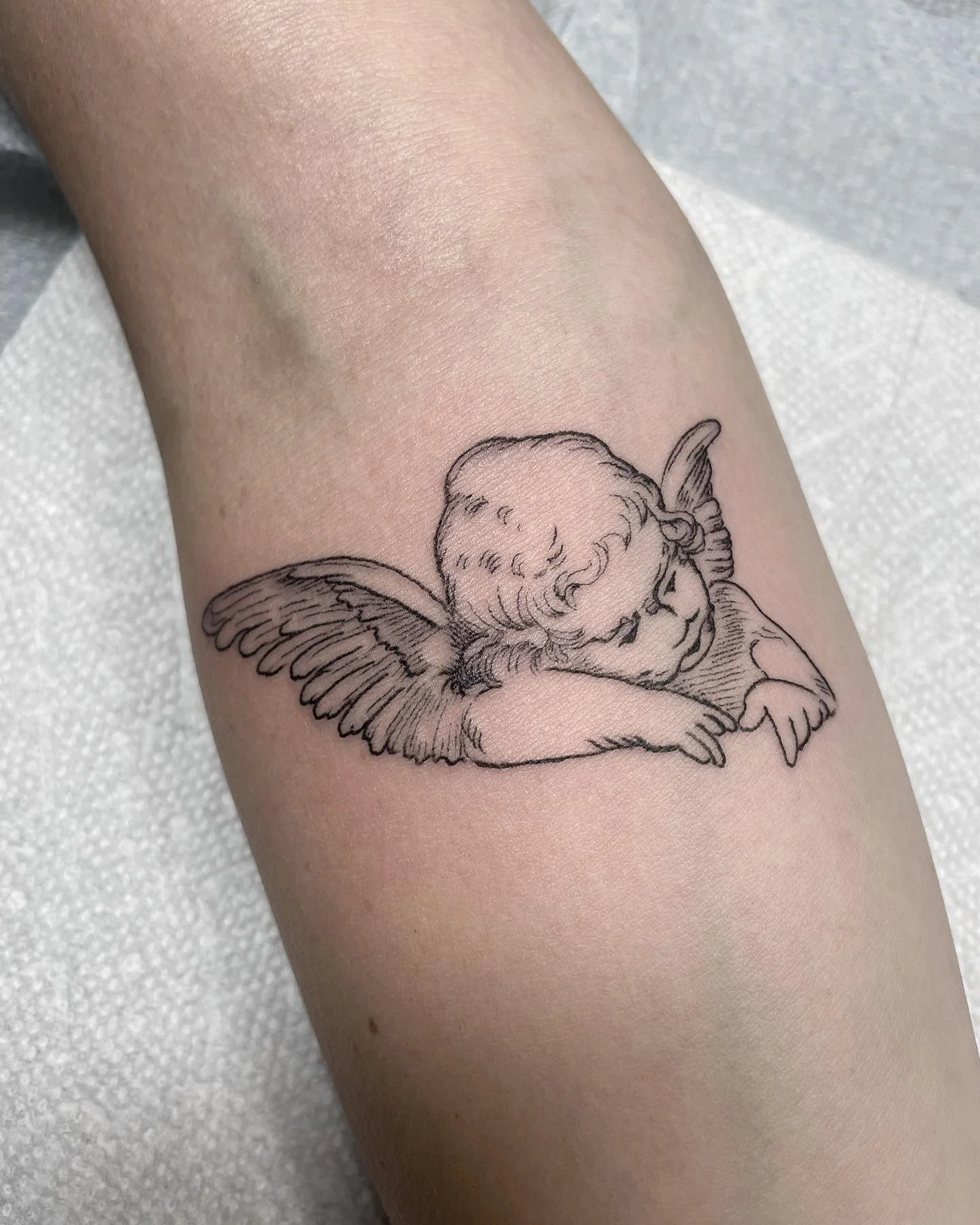 Line art tattoo of a cherub with wings resting its head on crossed arms.