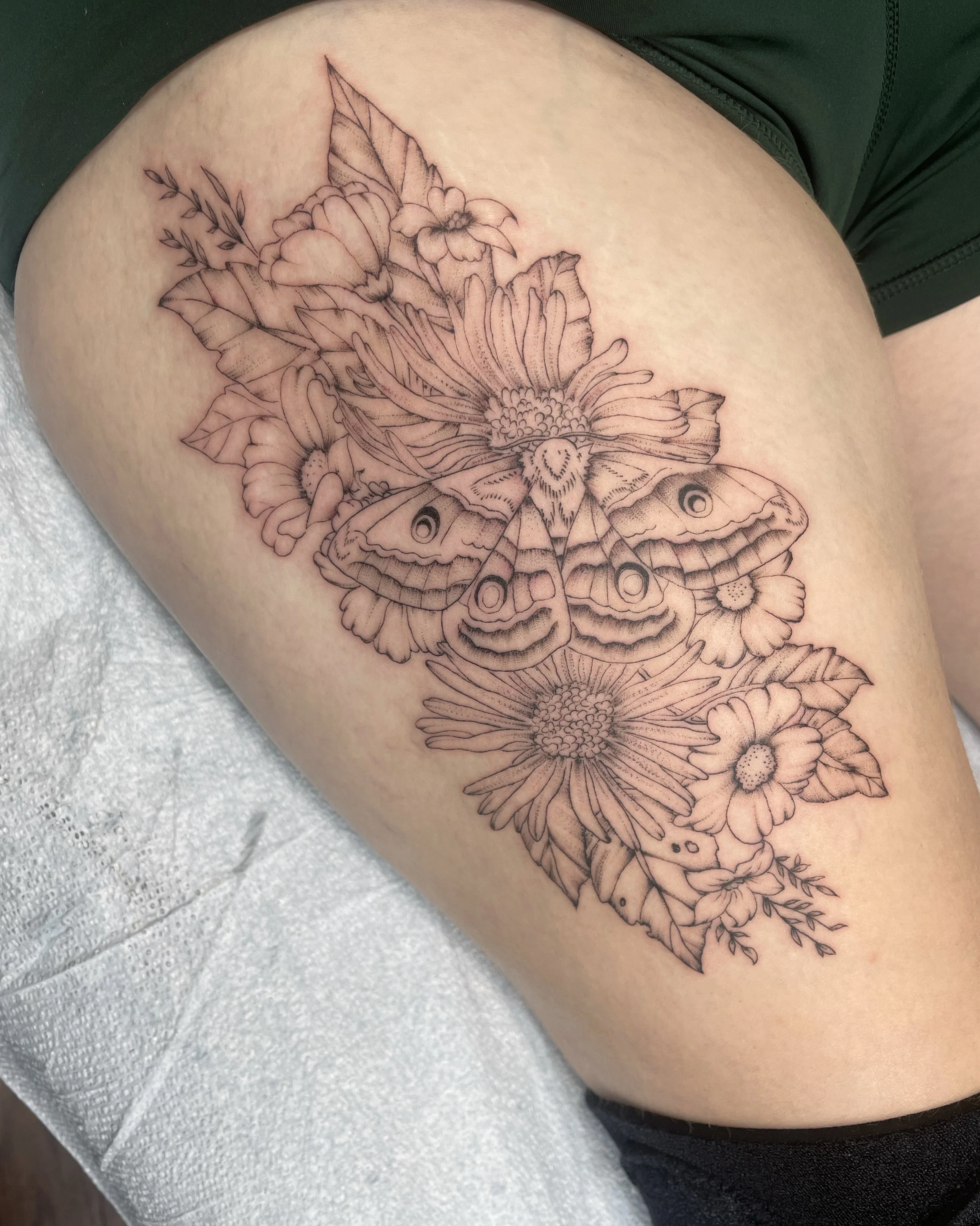 Line art tattoo of a butterfly surrounded by flowers and leaves on a person's thigh.