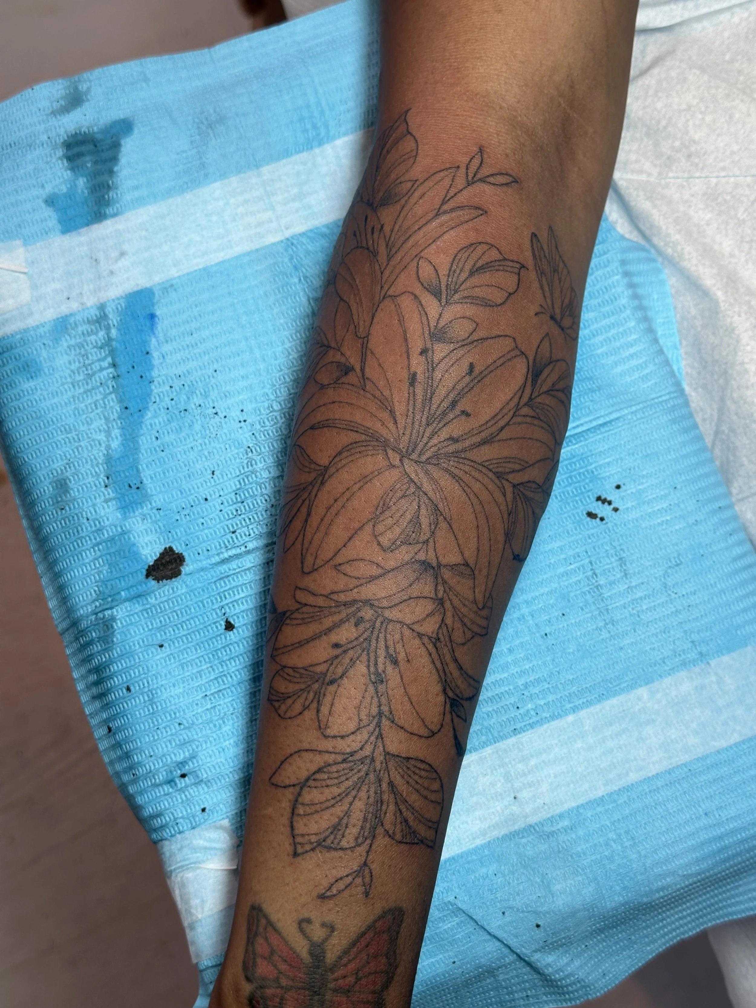 A tattoo of a large floral design with multiple leaves and petals on a person's forearm, resting on a plastic-covered surface with ink stains.