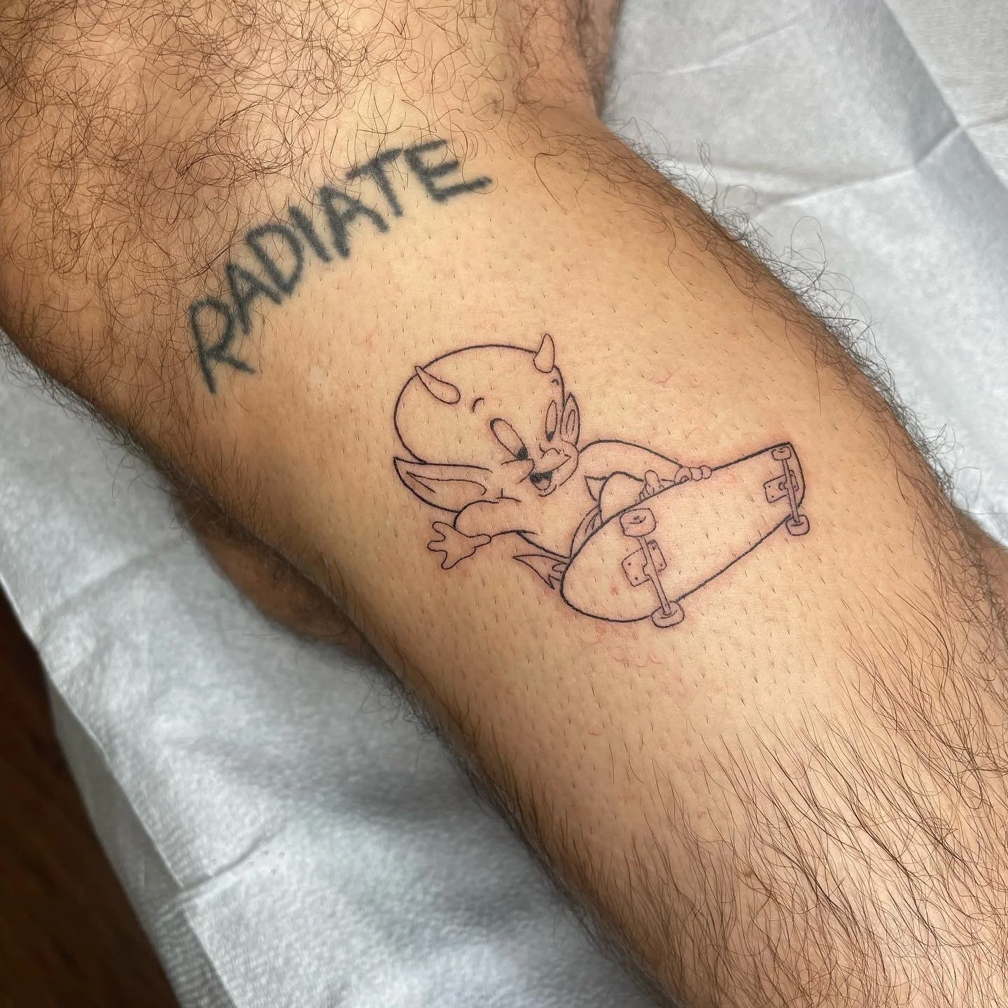 Tattoo of a cartoon devil riding a skateboard, positioned on the person's thigh. The devil has horns, wings, and a playful expression.