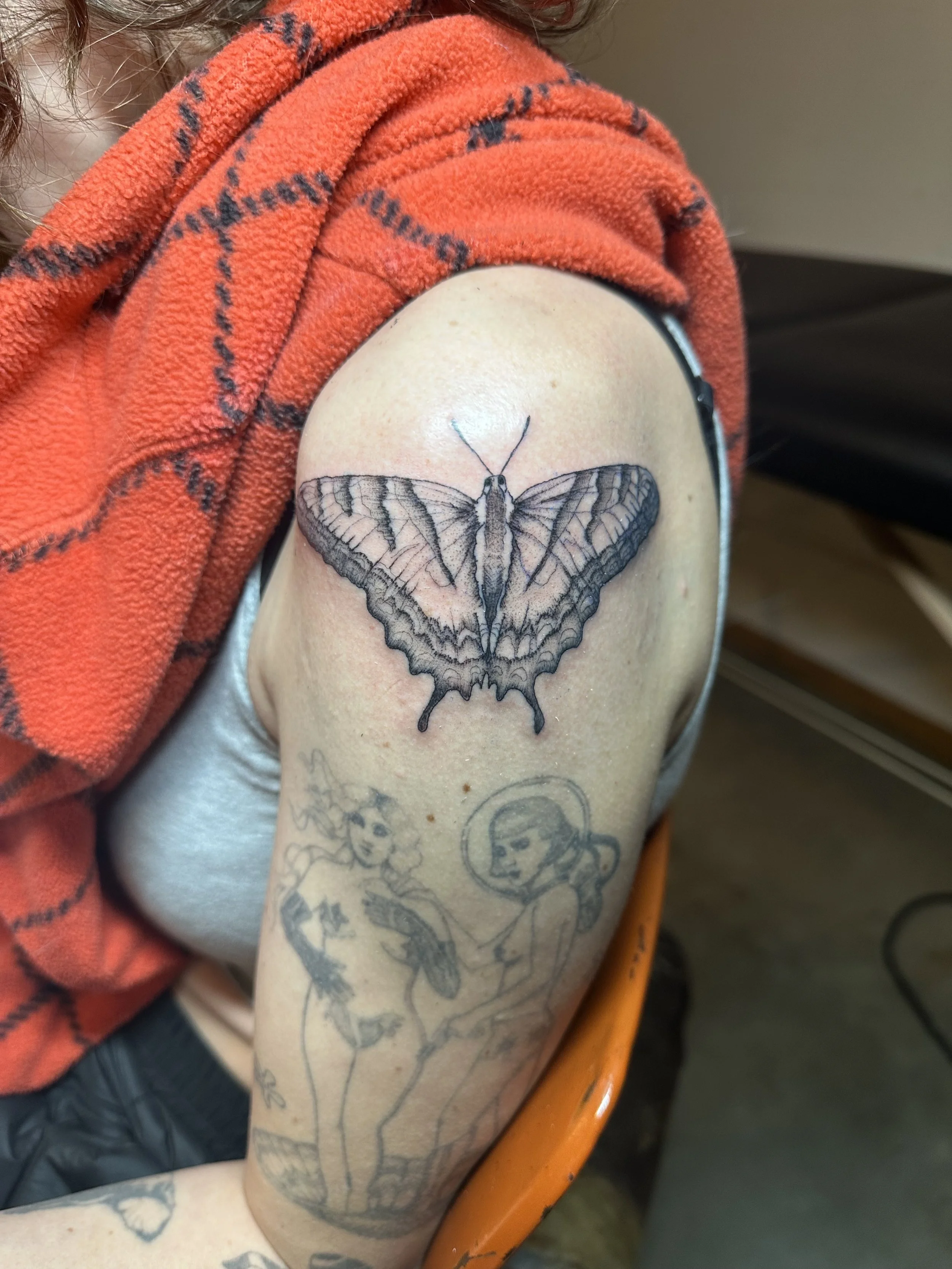 Tattoo of a butterfly on the upper arm, with a sketch of two women beneath it, one with curly hair and a flower, the other with a halo and a space helmet.