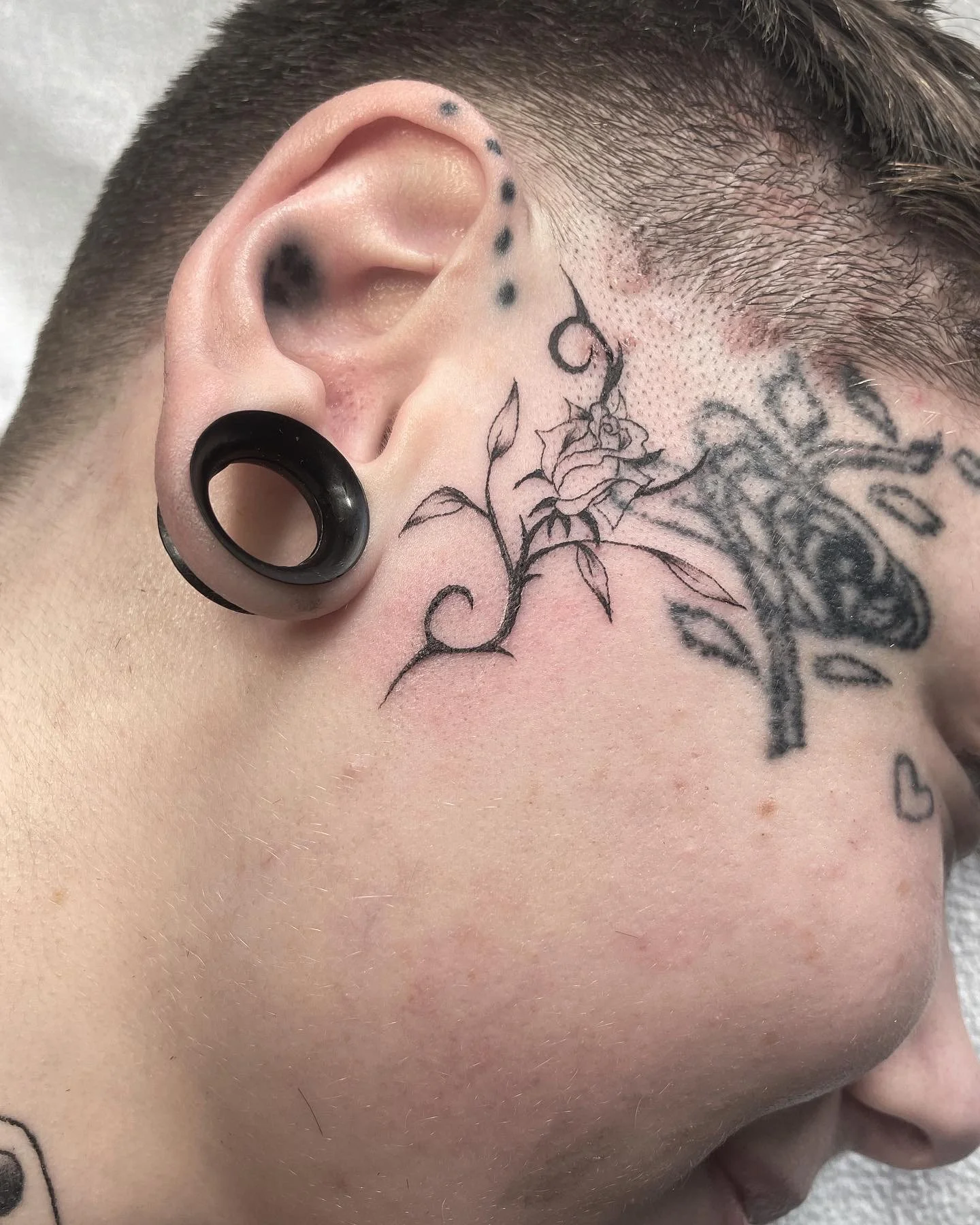 Close-up of a person's face showing a tattoo of a rose and vine next to the ear, with black ear gauges and multiple small dots in the hairline.
