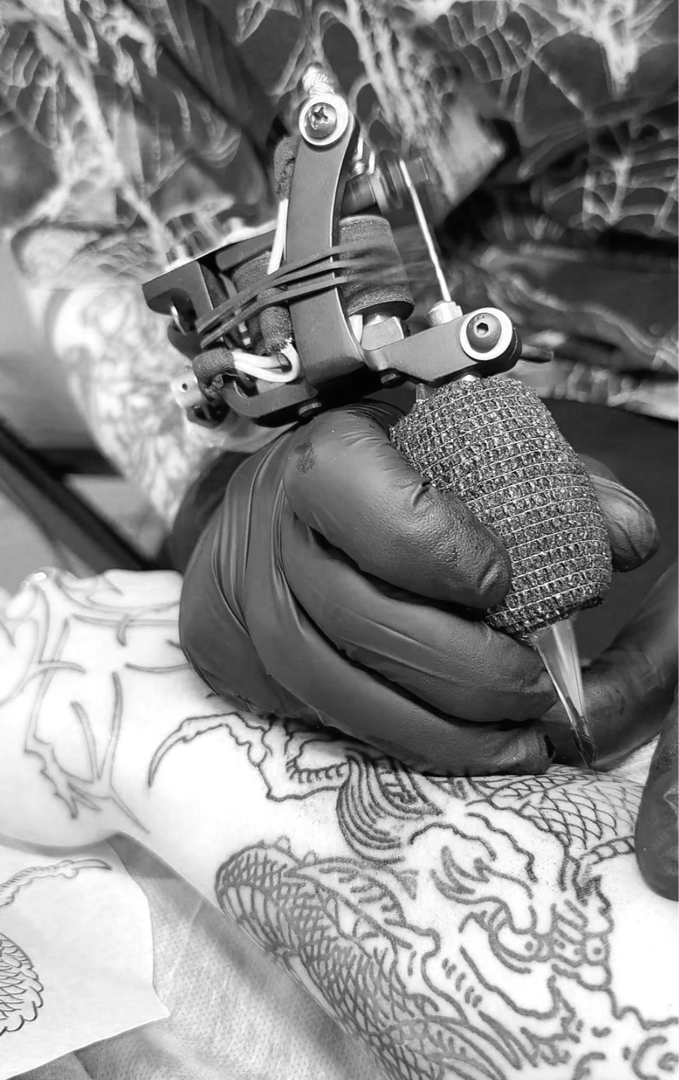 A tattoo artist's hand wearing a black glove, holding a tattoo machine and tattooing a dragon design on a person's arm.