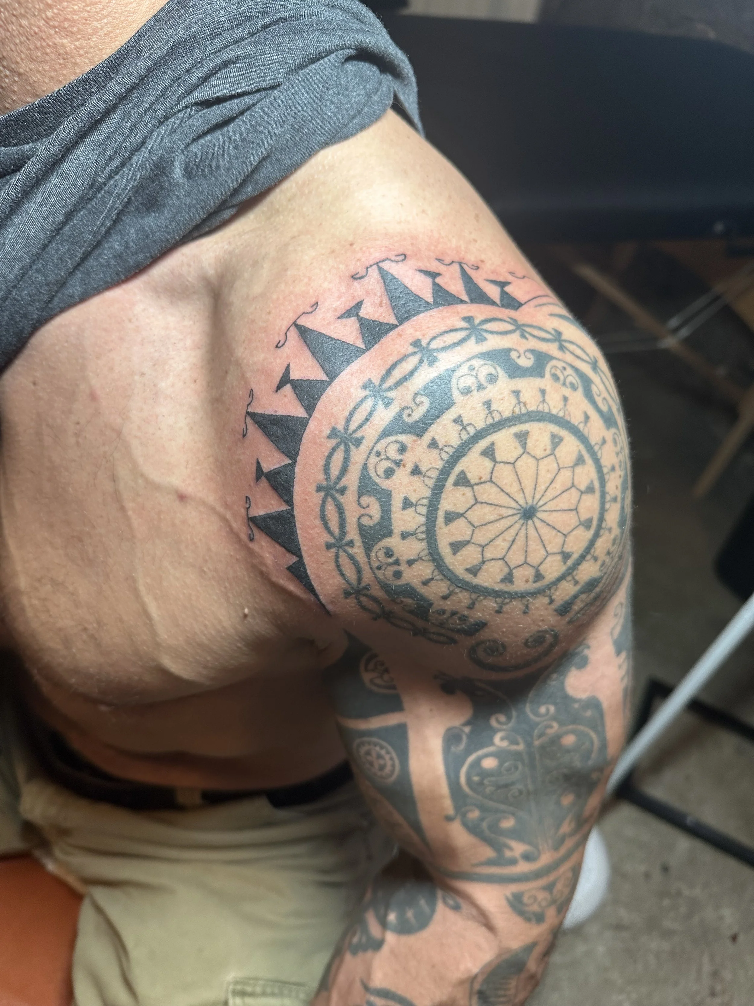 Close-up of a person's shoulder and arm with a large, intricate black and gray tattoo featuring geometric patterns and floral motifs.