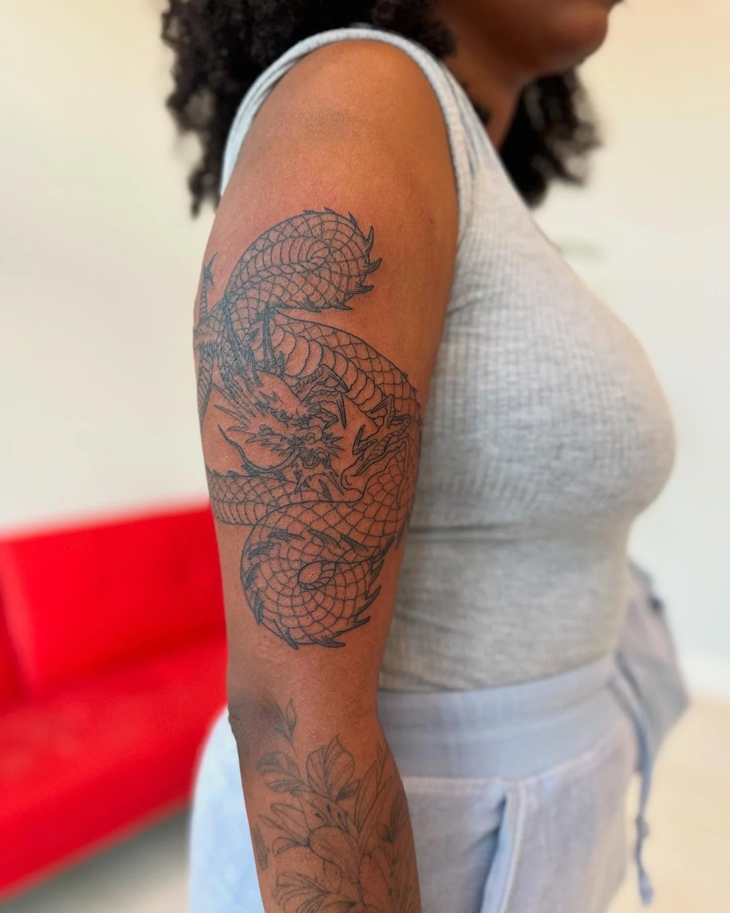 A woman with dark hair and skin is showing her upper arm tattoo of a dragon. She is wearing a light gray sleeveless top and light gray sweatpants with a drawstring. The background has a red chair and a light-colored wall.