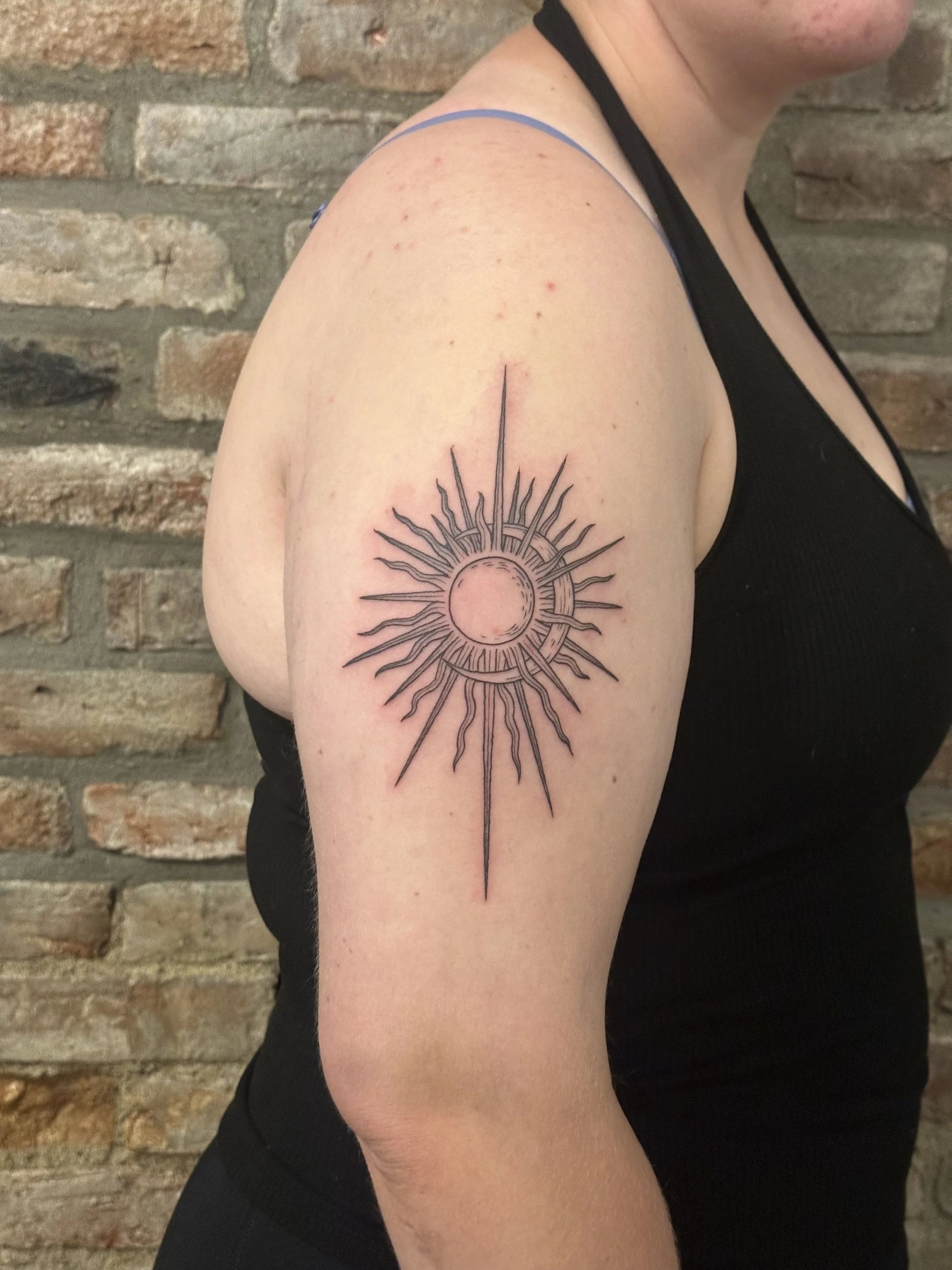 Tattoo of a sun with a circle in the center and radiating rays on the upper arm of a person wearing a black sleeveless top.