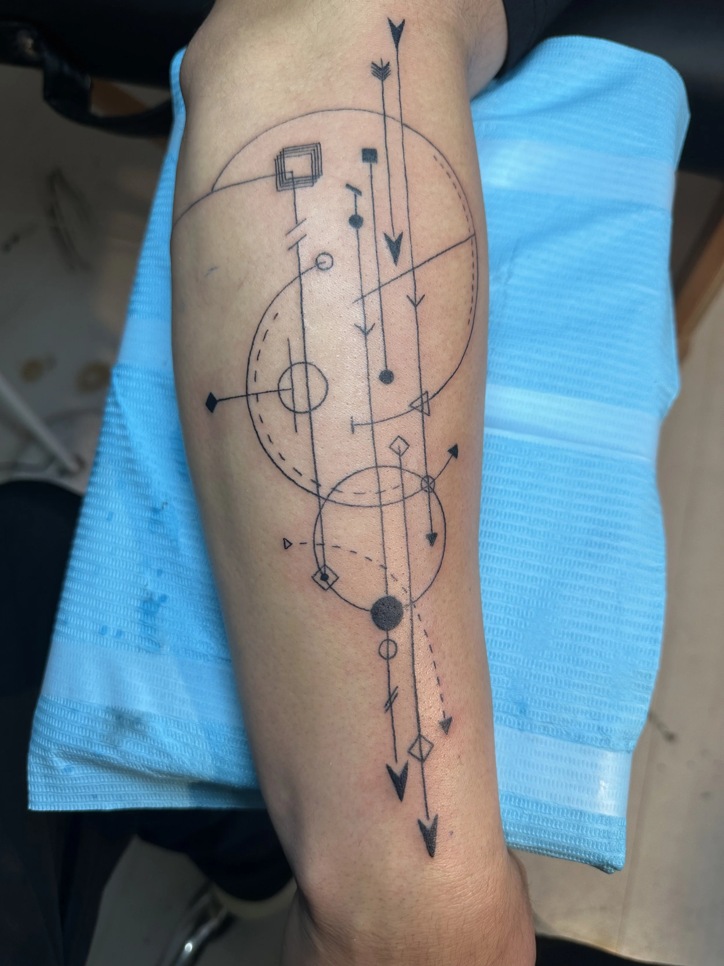 A tattoo on a person's forearm featuring a geometric and abstract design with various circles, lines, arrows, and symbols in black ink.