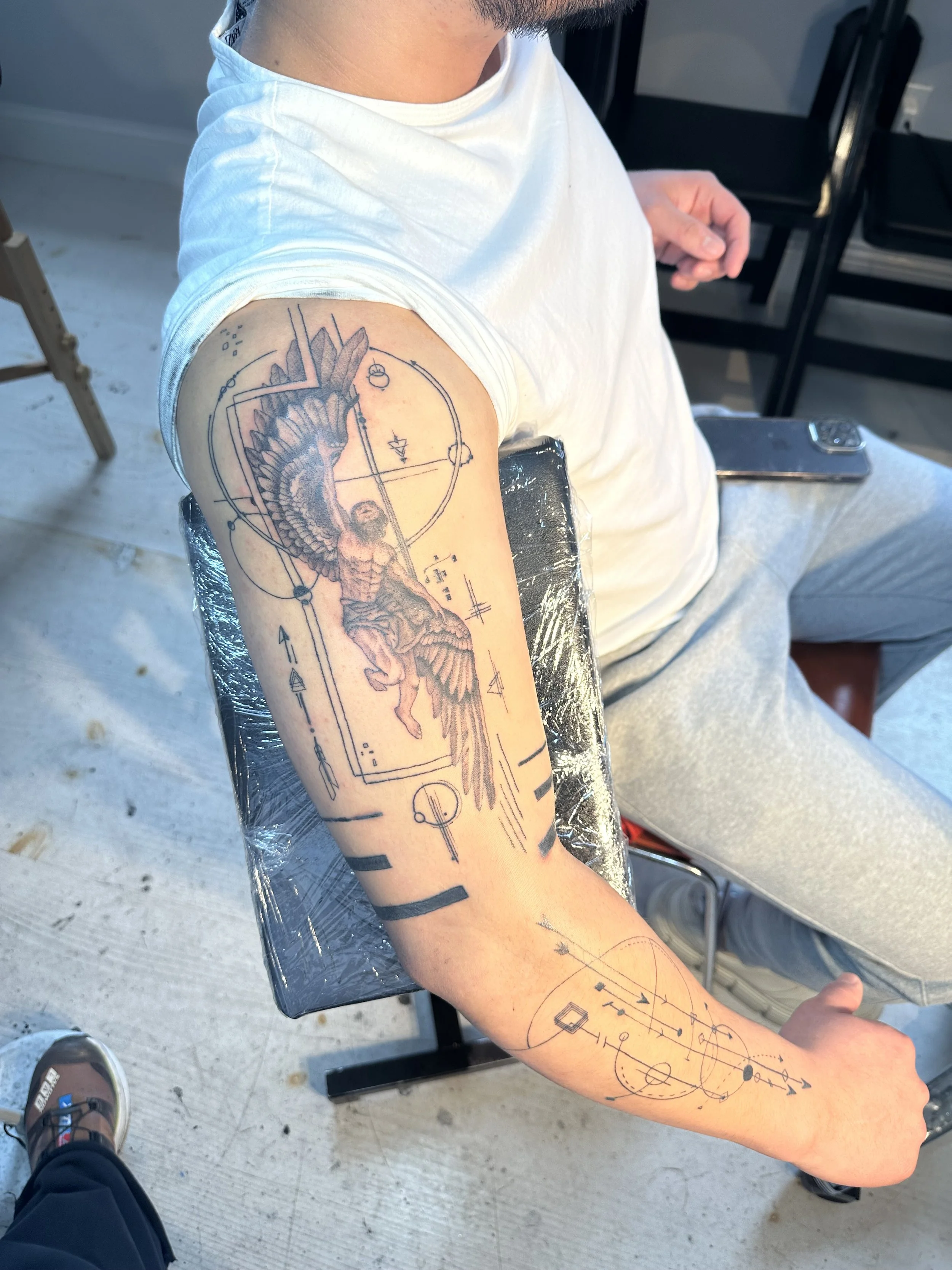 A person sitting in a chair with a tattoo design in progress on their upper arm. The tattoo depicts a figure with wings and various geometric shapes and lines surrounding it.