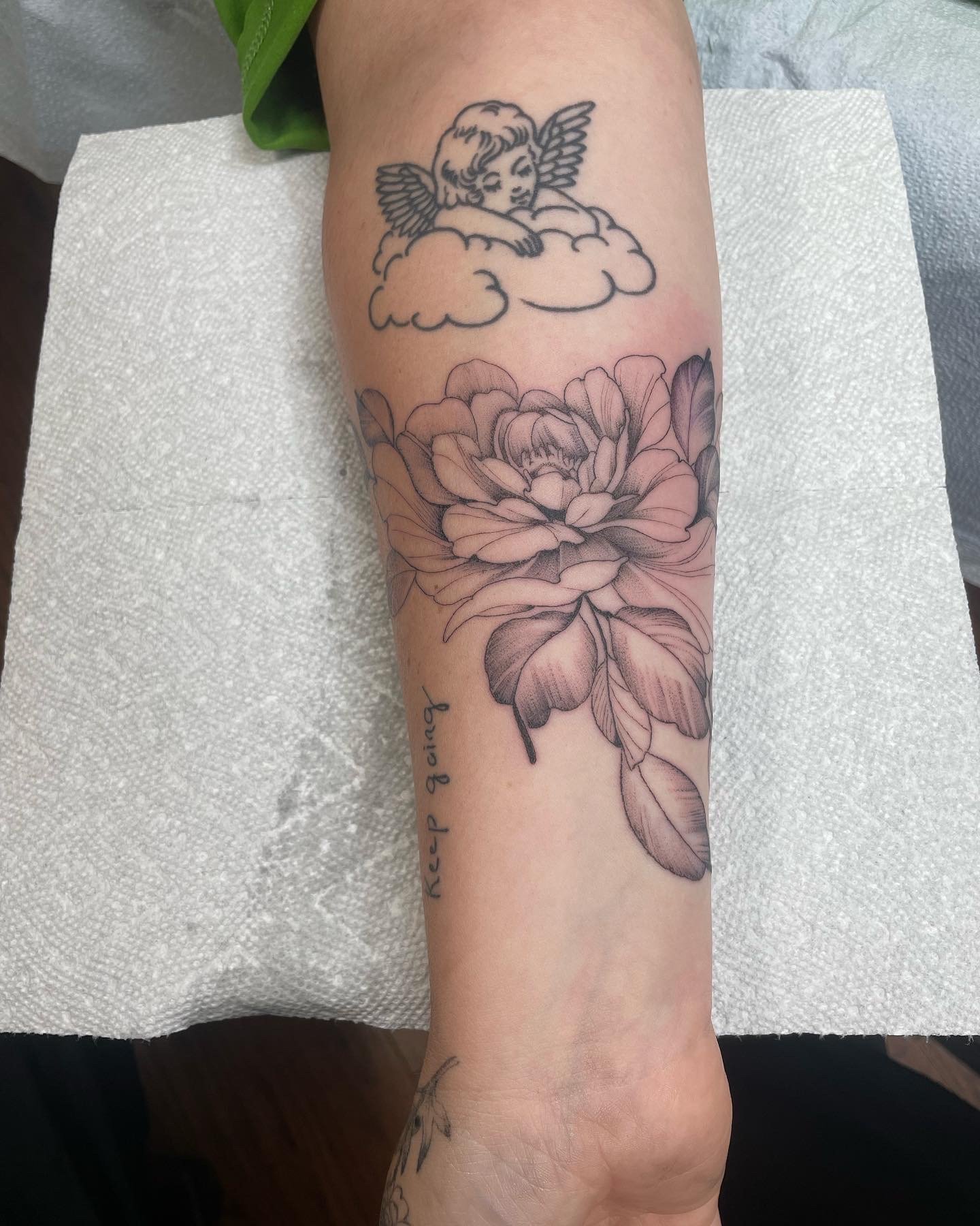 Tattoo of a cherub with wings resting on a cloud above a large detailed peony flower with leaves, and a tattooed phrase 'keep a faith' on the wrist.
