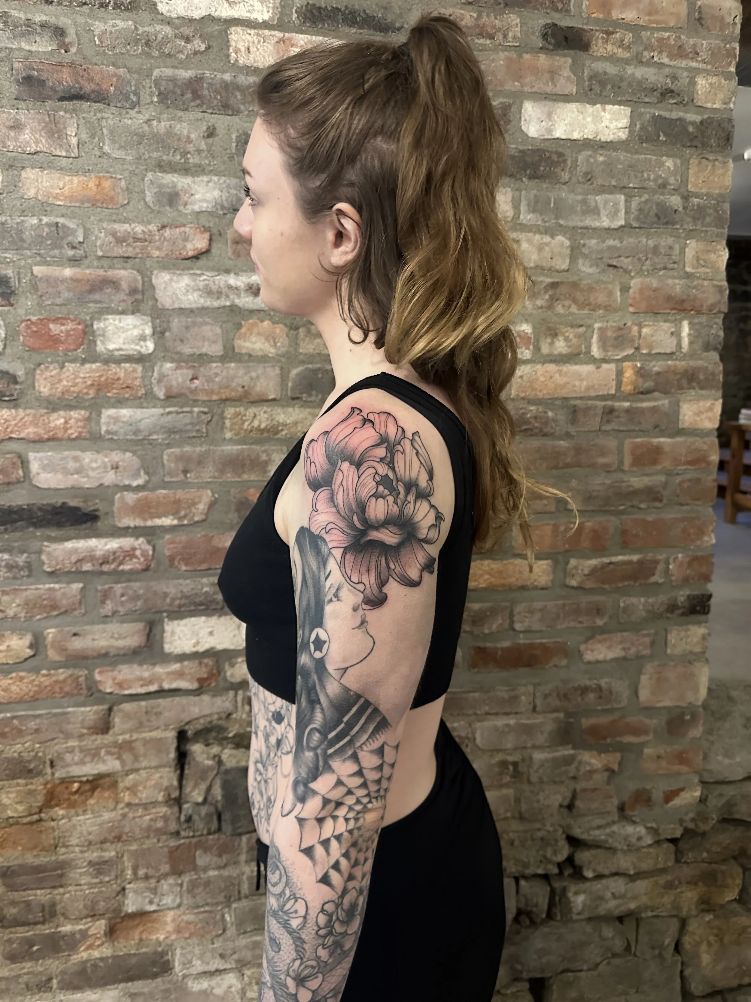 A woman with wavy, shoulder-length brown hair standing sideways against a brick wall, wearing a black sleeveless top. She has a large, detailed tattoo featuring a woman's profile, large flower, and geometric patterns on her upper arm.