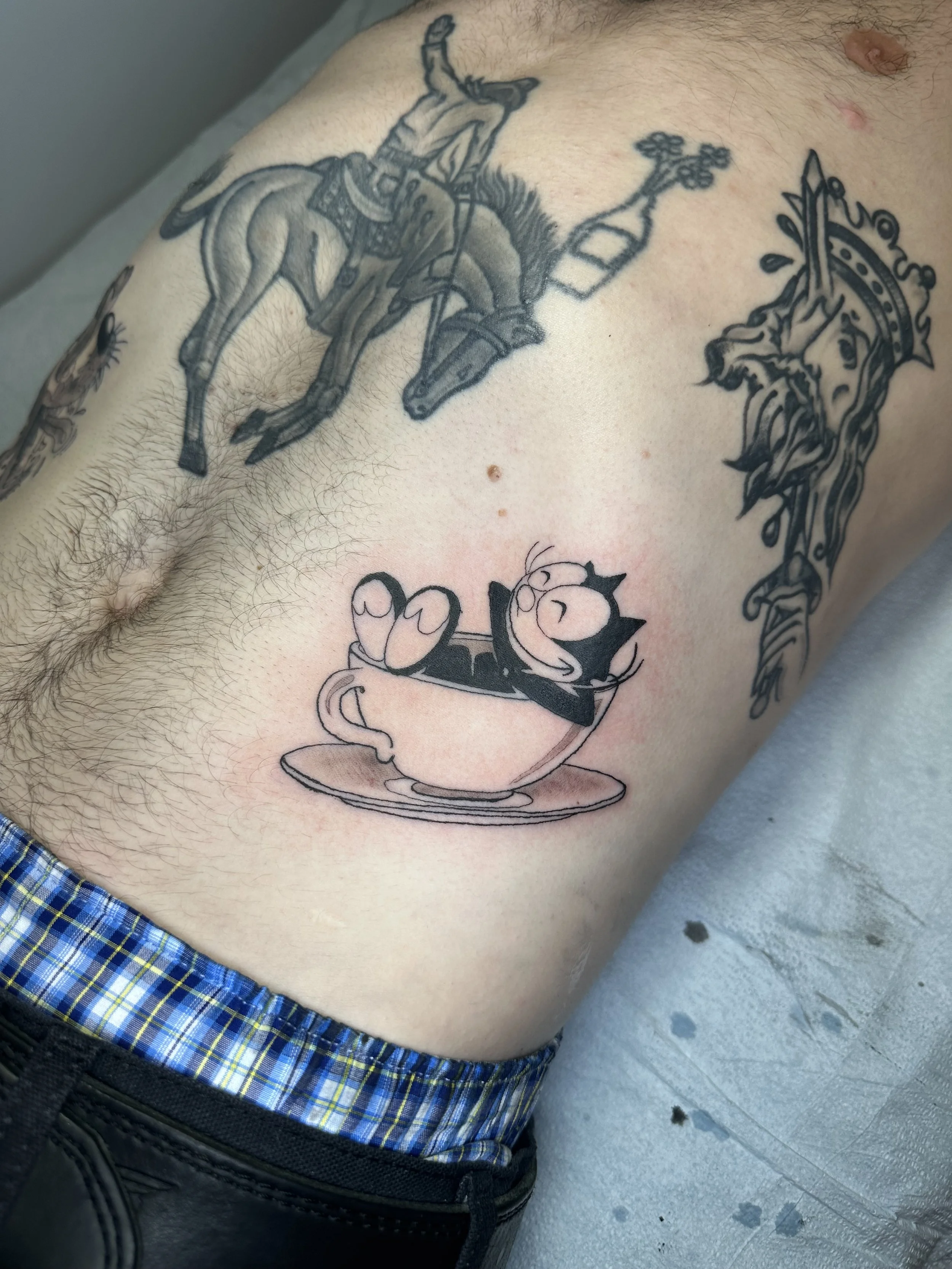 Tattoos on a person's side, including a cartoon cat sleeping in a cup and a detailed knight riding a horse with a lance.