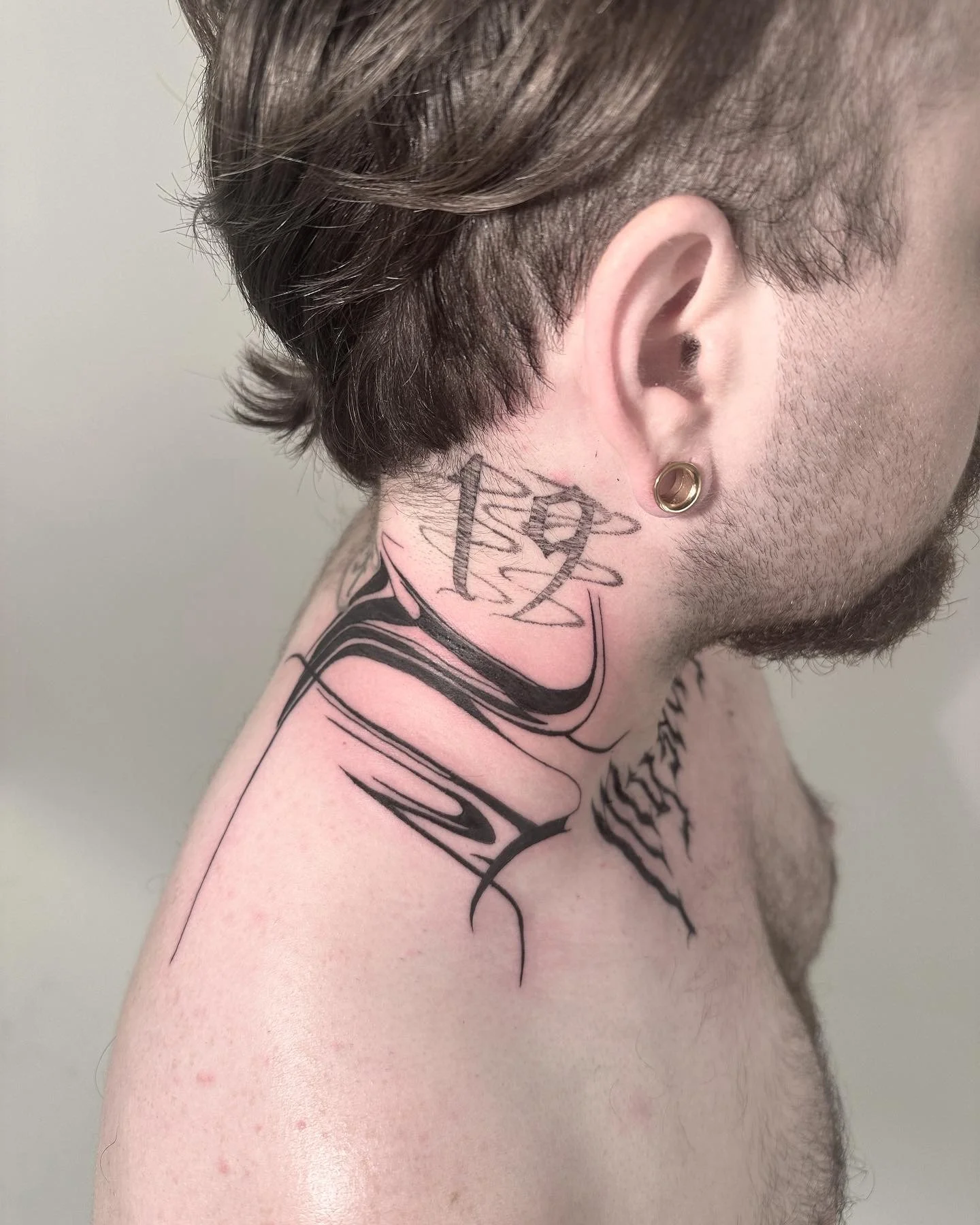 A person with short brown hair and a beard, showing side profile with a large tattoo on the neck and shoulder area. The tattoo includes a stylized, abstract design and the number '19' written over a winged clock face.