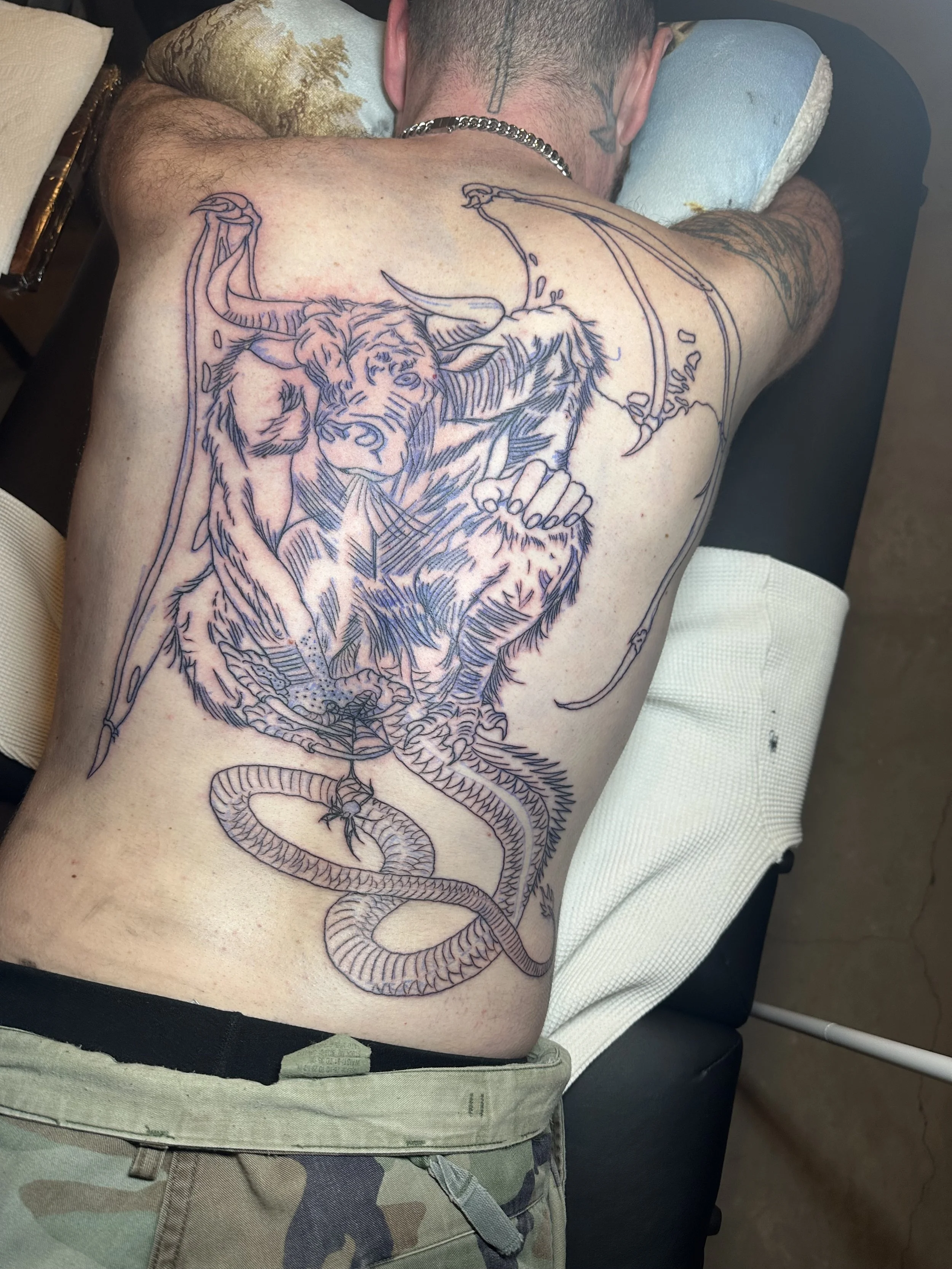 A man lying face down on a tattoo table, receiving a large tattoo of a tiger, snake, and umbrella on his back.