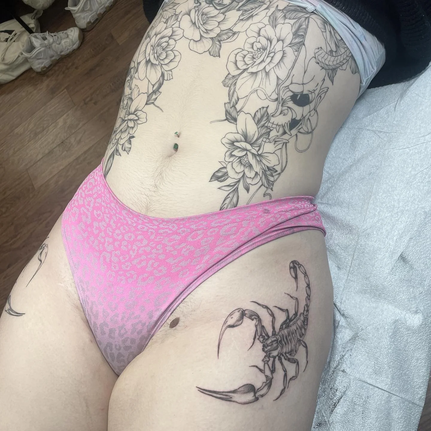 A person lying on a bed with tattoos, including a large floral design on the torso and a scorpion tattoo on the thigh, wearing pink leopard-print underwear.