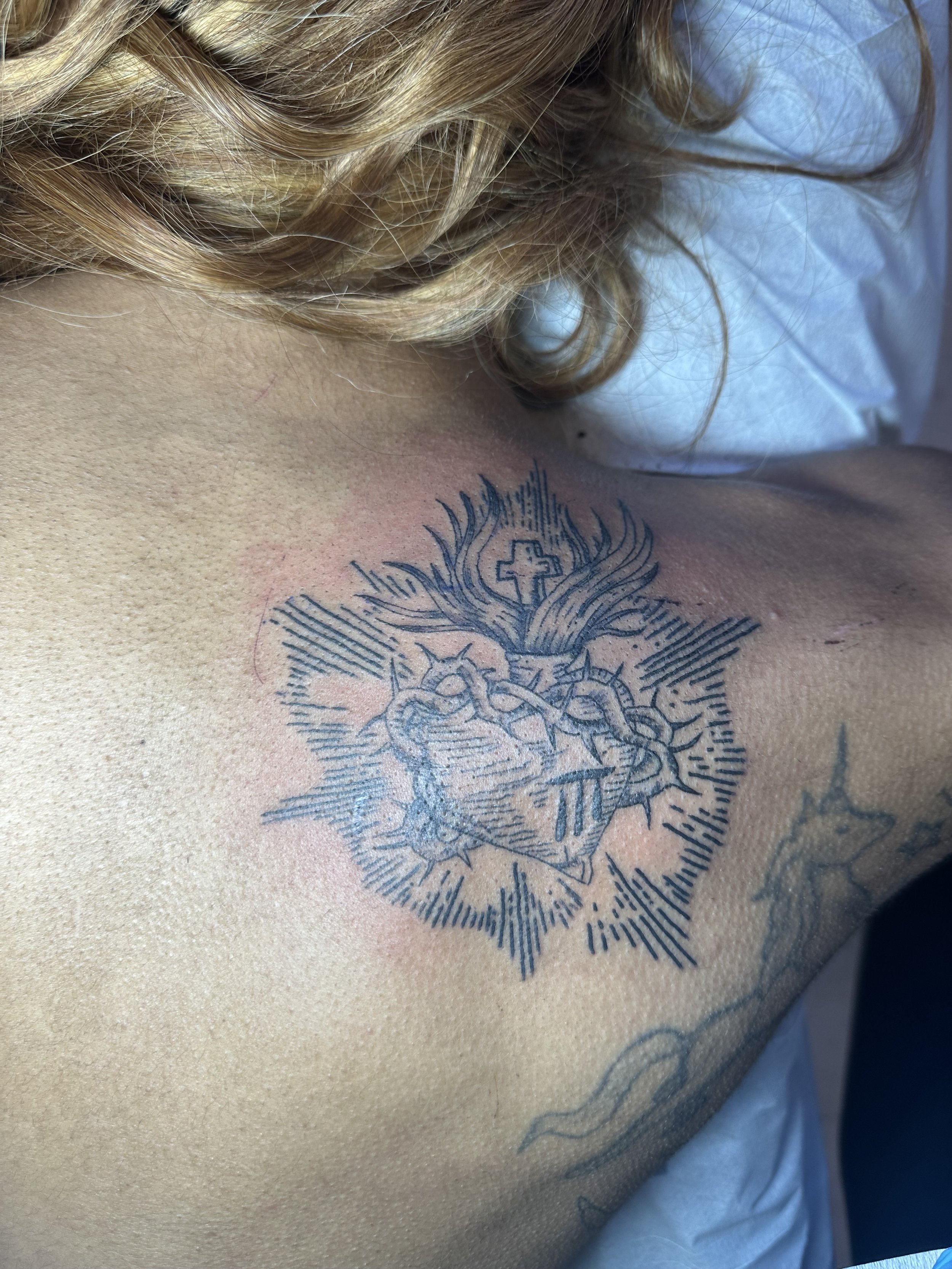 Tattoo of a heart with a cross, wings, and what appears to be a crown on the chest, on a person's upper chest near the collarbone.