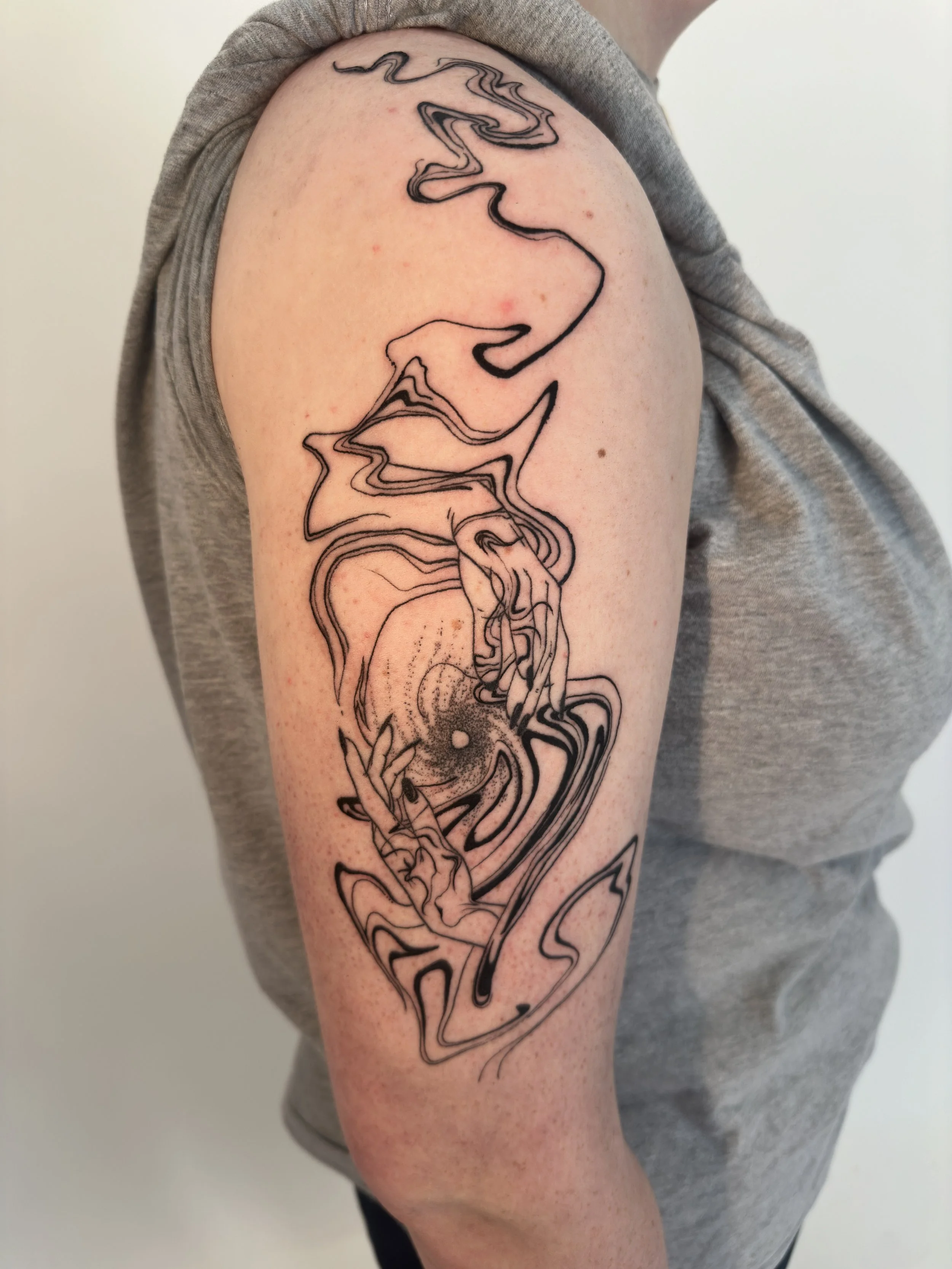 A black abstract line art tattoo on a person's upper arm, featuring swirling, fluid shapes and a central dot, with the person wearing a sleeveless grey shirt.