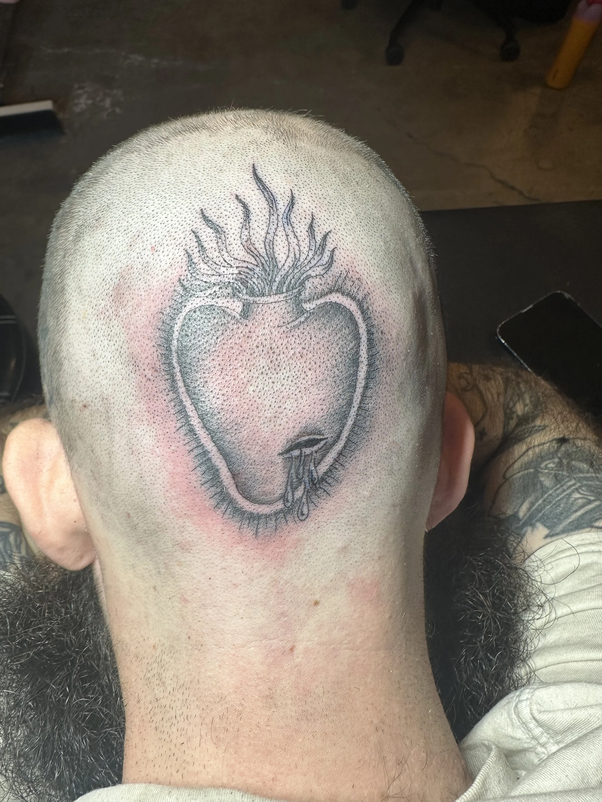 Tattoo of a heart with flames and tear drops on the back of a person's shaved head.