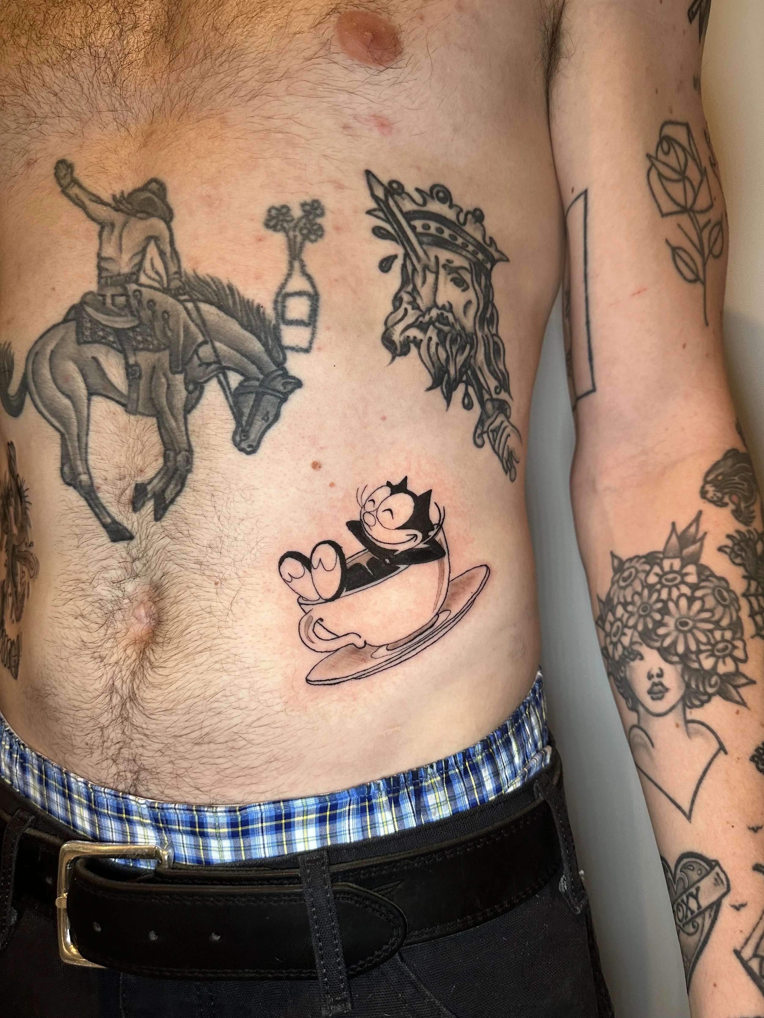 Tattooed torso with various tattoos, including a cartoon character in a spaceship, a black and gray depiction of Jesus with a crown, and a detailed sole flower tattoo on the arm.