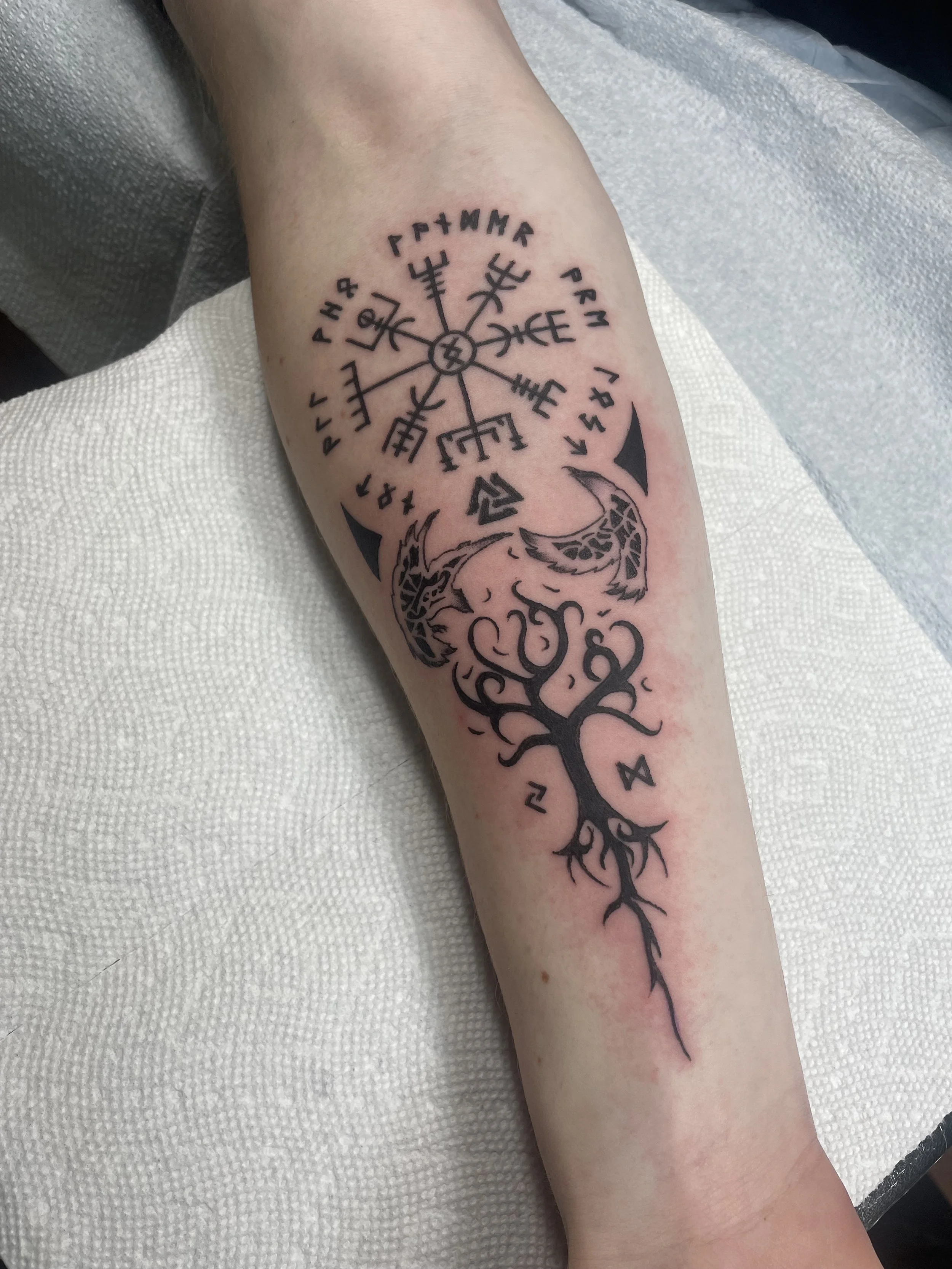 A tattoo on a person's forearm featuring a tree with branches and roots, a series of runes and symbols, and a circular design at the top with intersecting lines and symbols.