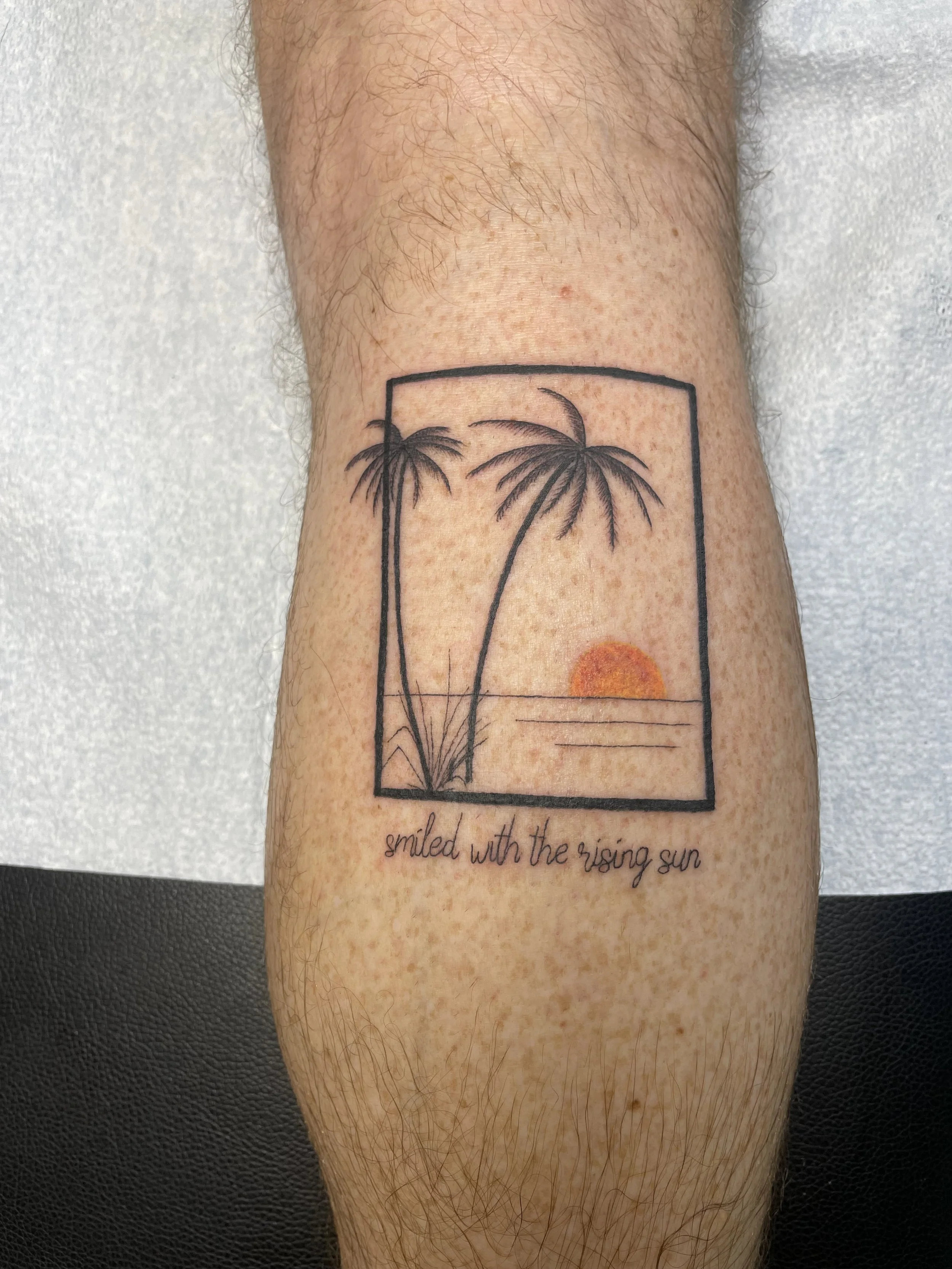 Tattoo of two palm trees and a sunset within a framed square, with the words "smiled with the rising sun" written below.