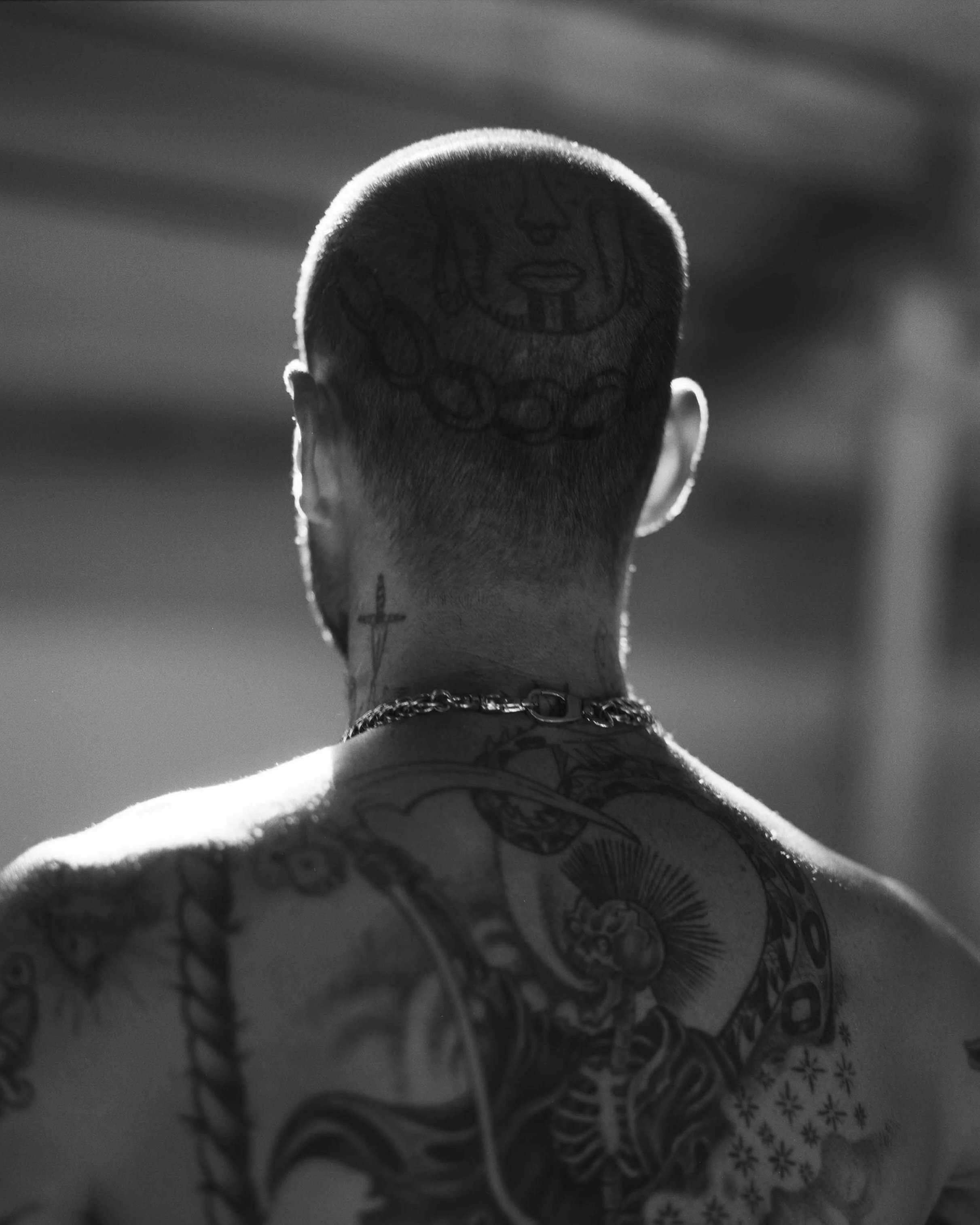 Back view of a tattooed man with a shaved head featuring a circular tattoo of a face surrounded by chains. He has tattoos on his shoulders and neck, and is wearing earrings and a chain necklace, with sunlight highlighting the outlines.