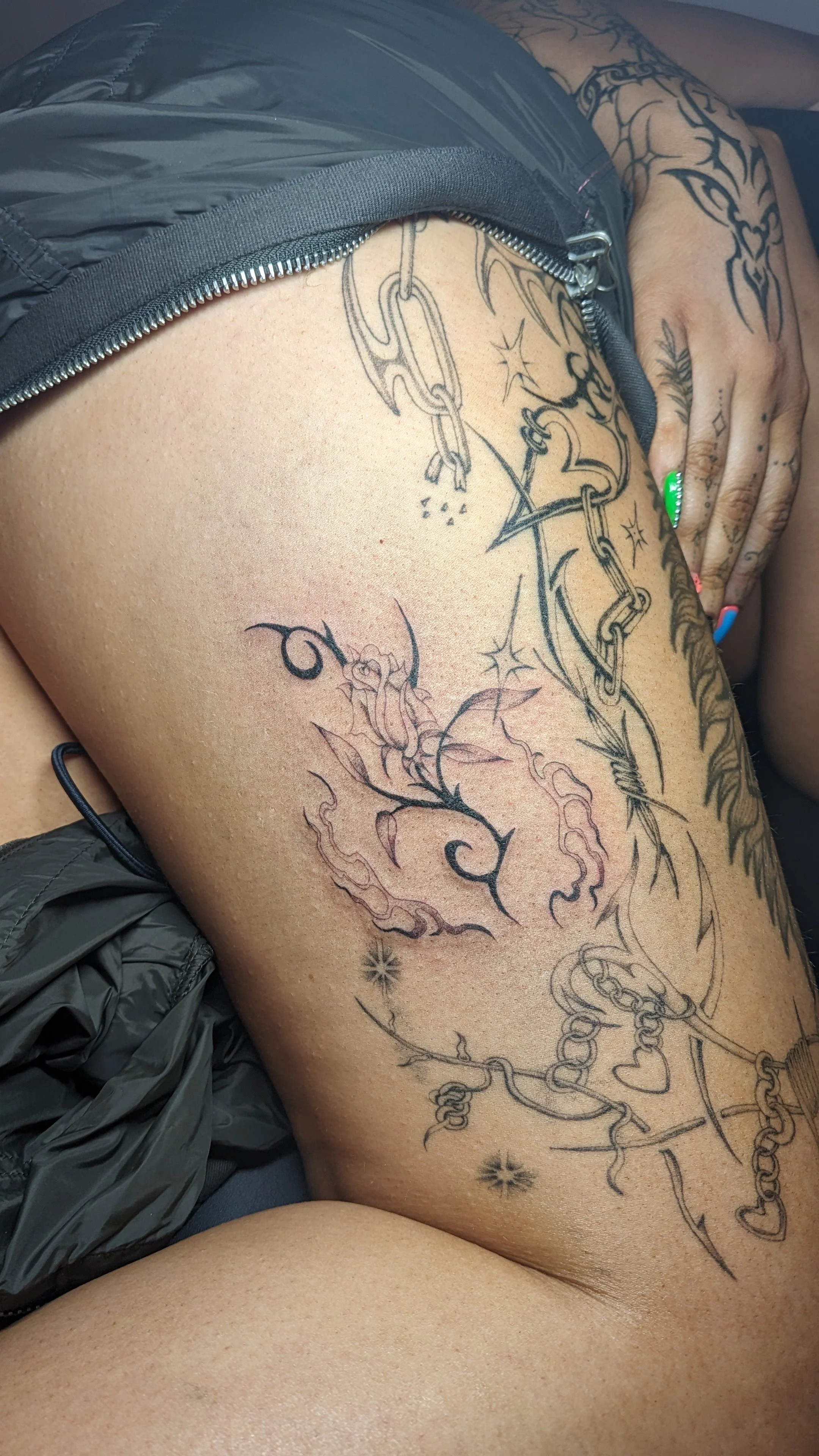 A person's thigh with various tattoos, including a dragon, chains, stars, and floral designs, partially covered by grey clothing.
