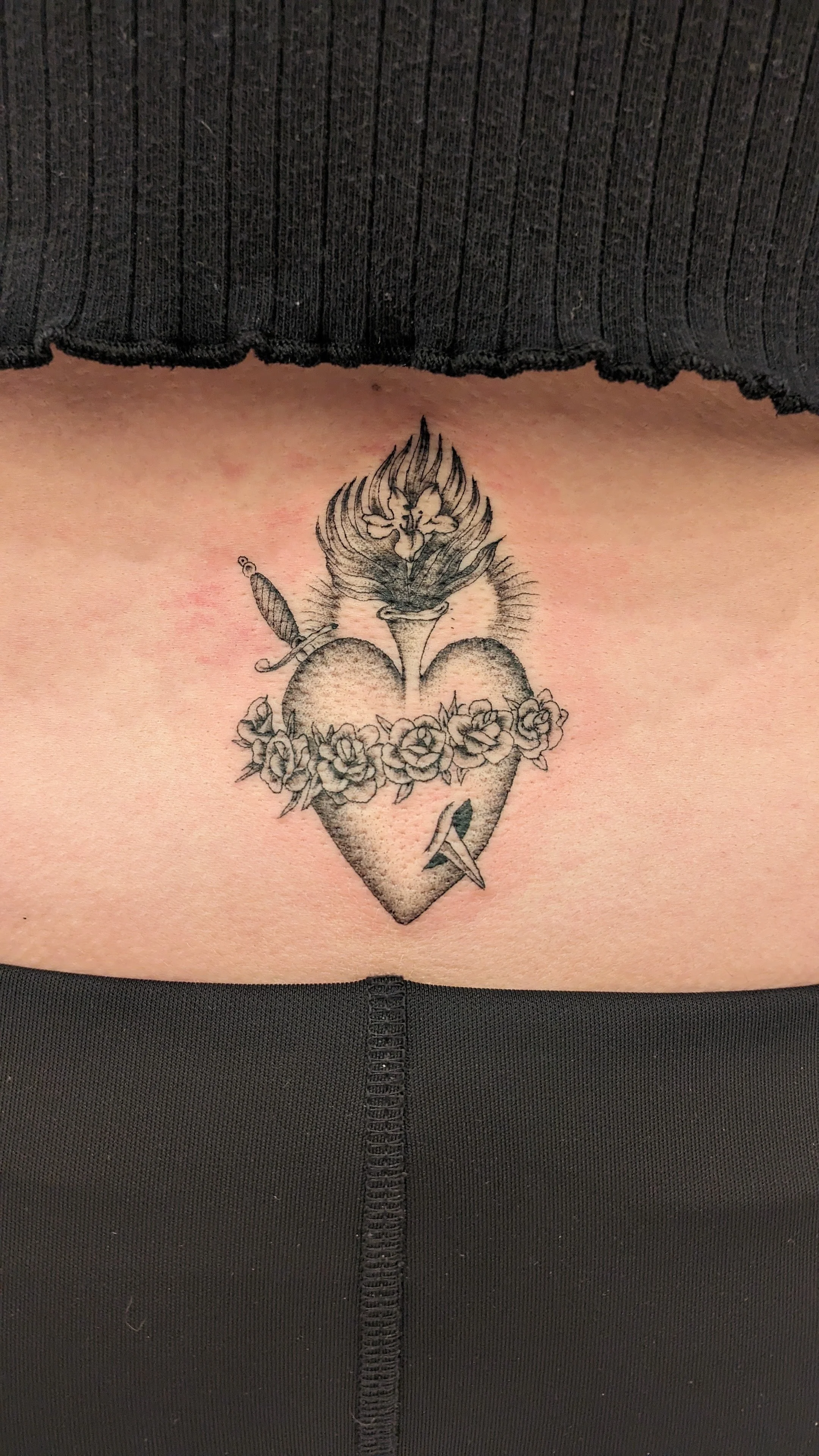 Tattoo of a heart with a flower crown, a dagger, and a flame at the top, located on someone's side.