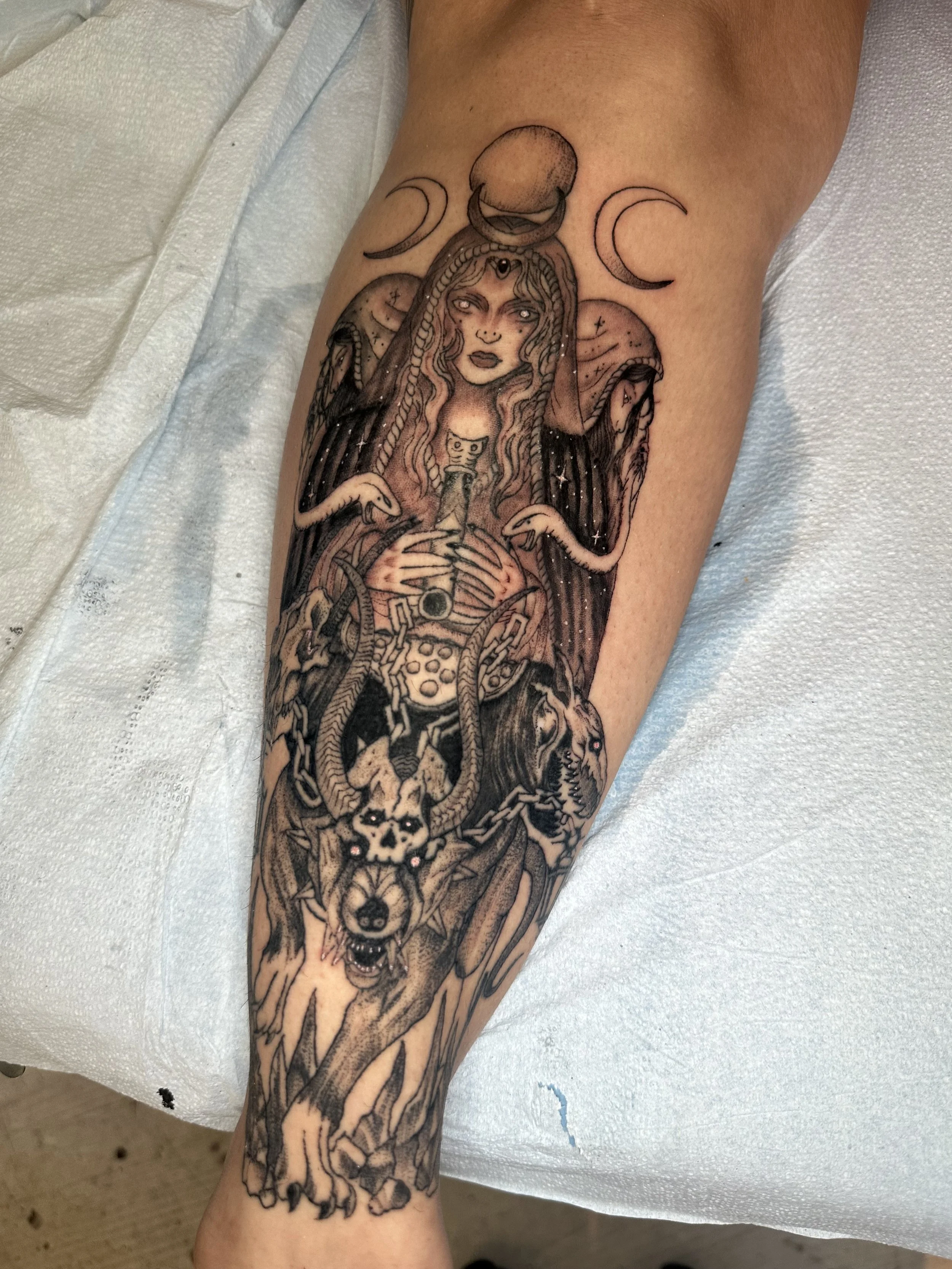 Tattoo of a mystical woman with long hair, holding a sword, with a crescent moon, a full moon, and two smaller moons above her head. She has goat skulls, wolves, and chains at the bottom, with starry details and a dark background.