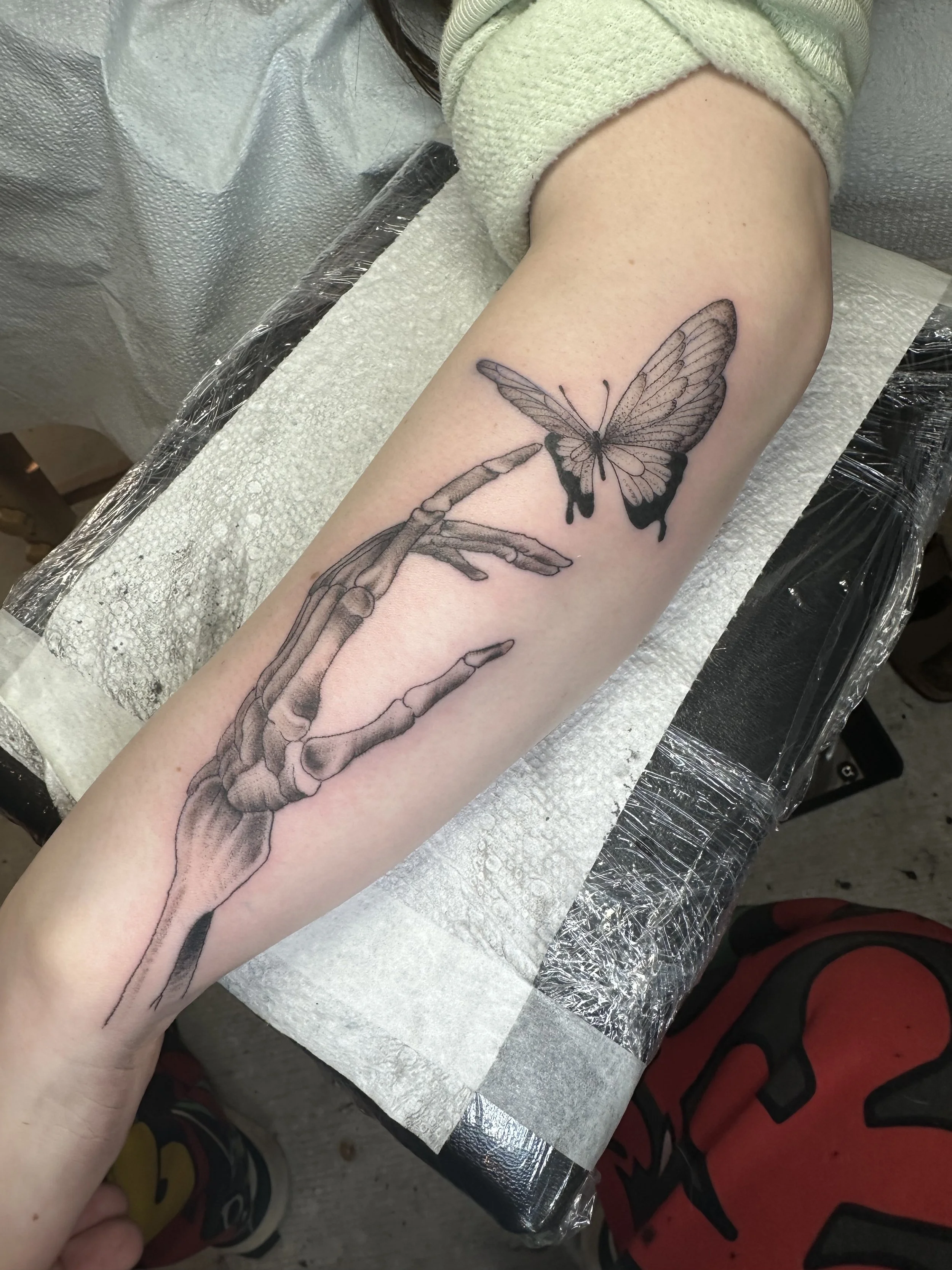 A tattoo of a butterfly and a skeleton hand on a person's arm, with the skeleton hand reaching towards the butterfly.