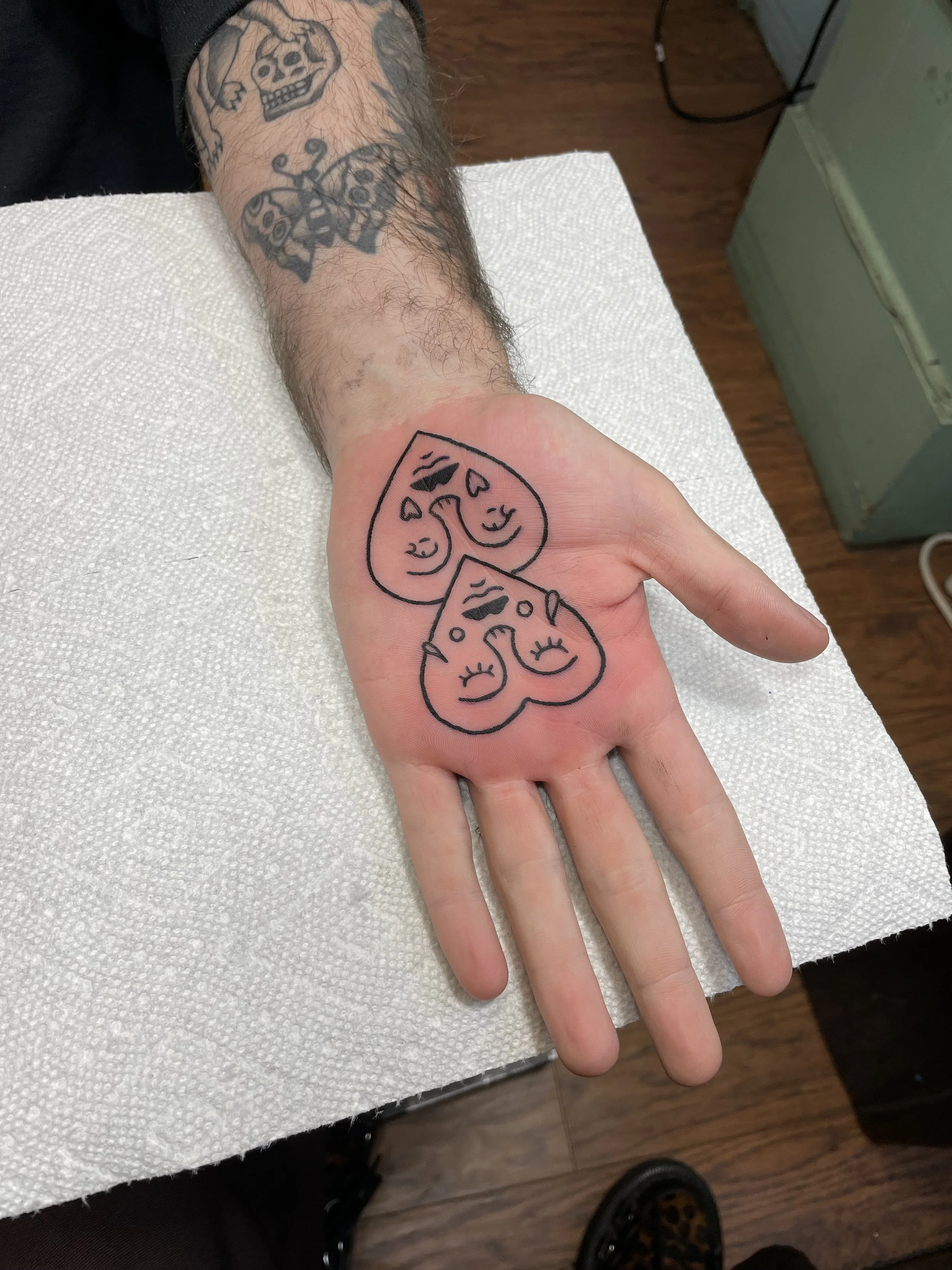 A person's hand with a new tattoo of two cartoon-like smiling faces with closed eyes and heart shapes above their heads, outlined in black ink.