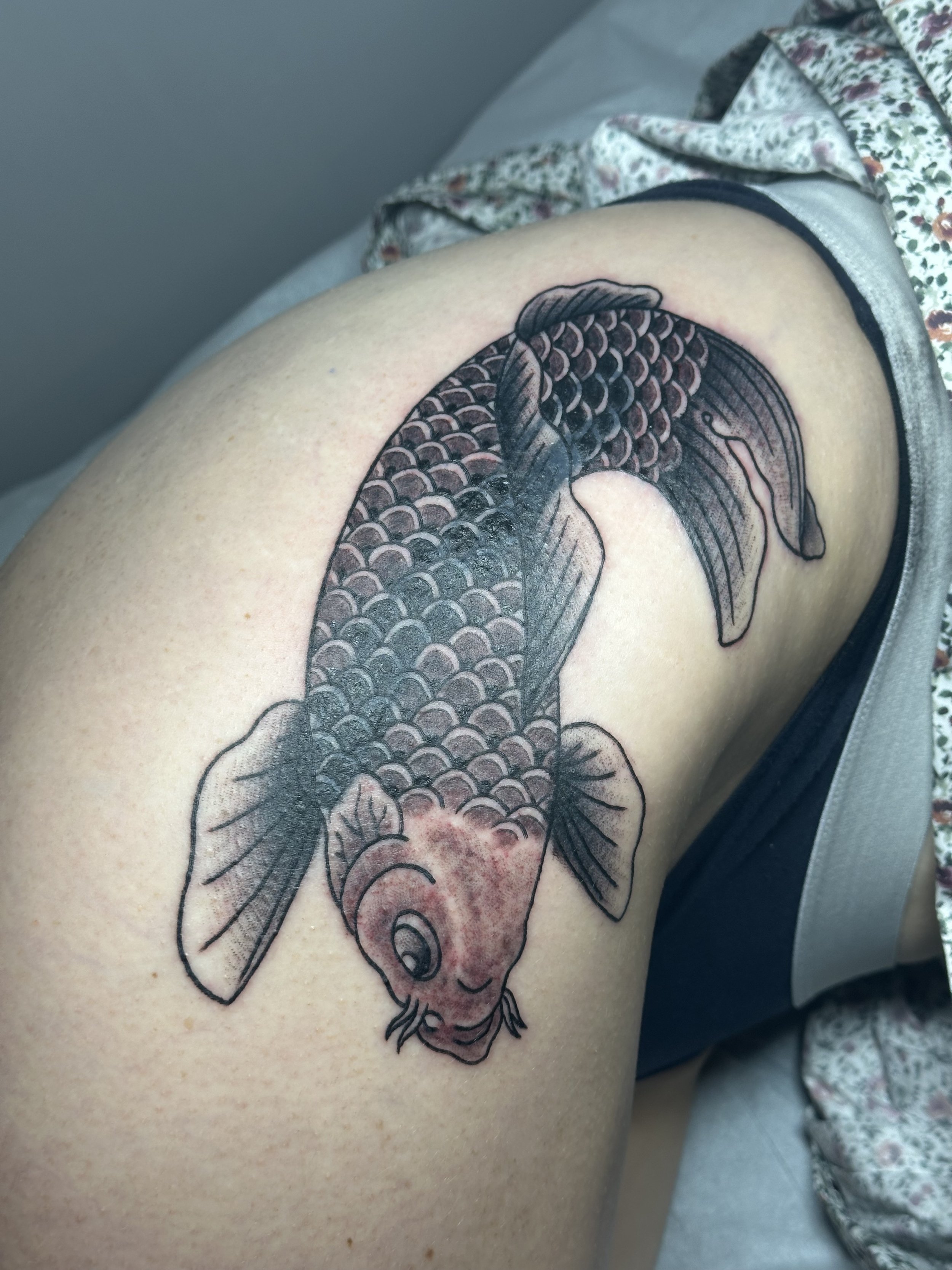 Tattoo of a koi fish on a person's thigh, featuring detailed scales, fins, and a detailed face, in black ink with shading.