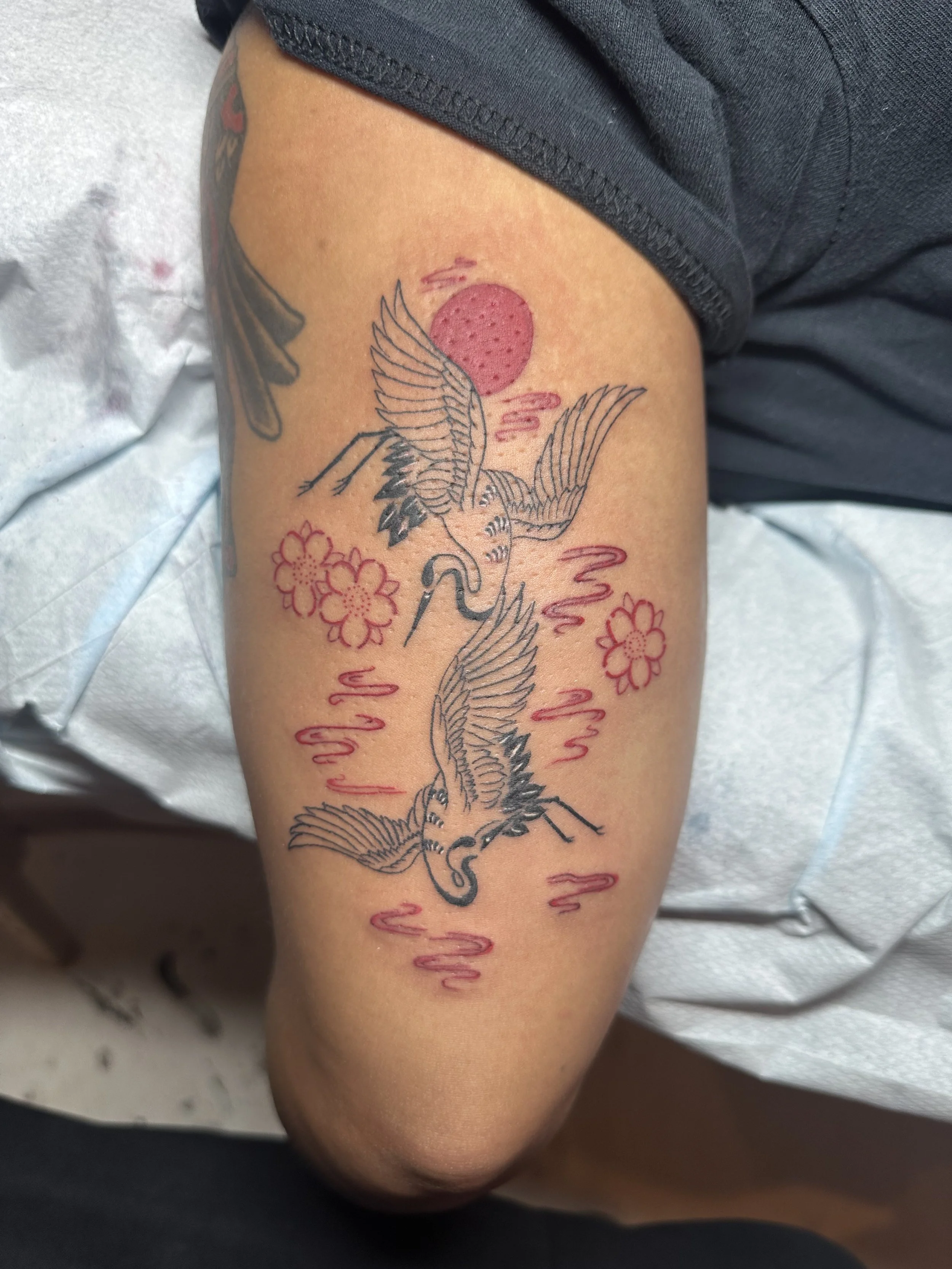 Tattoo of two cranes flying with pink flowers and a pink sun in the background on a person's upper arm.