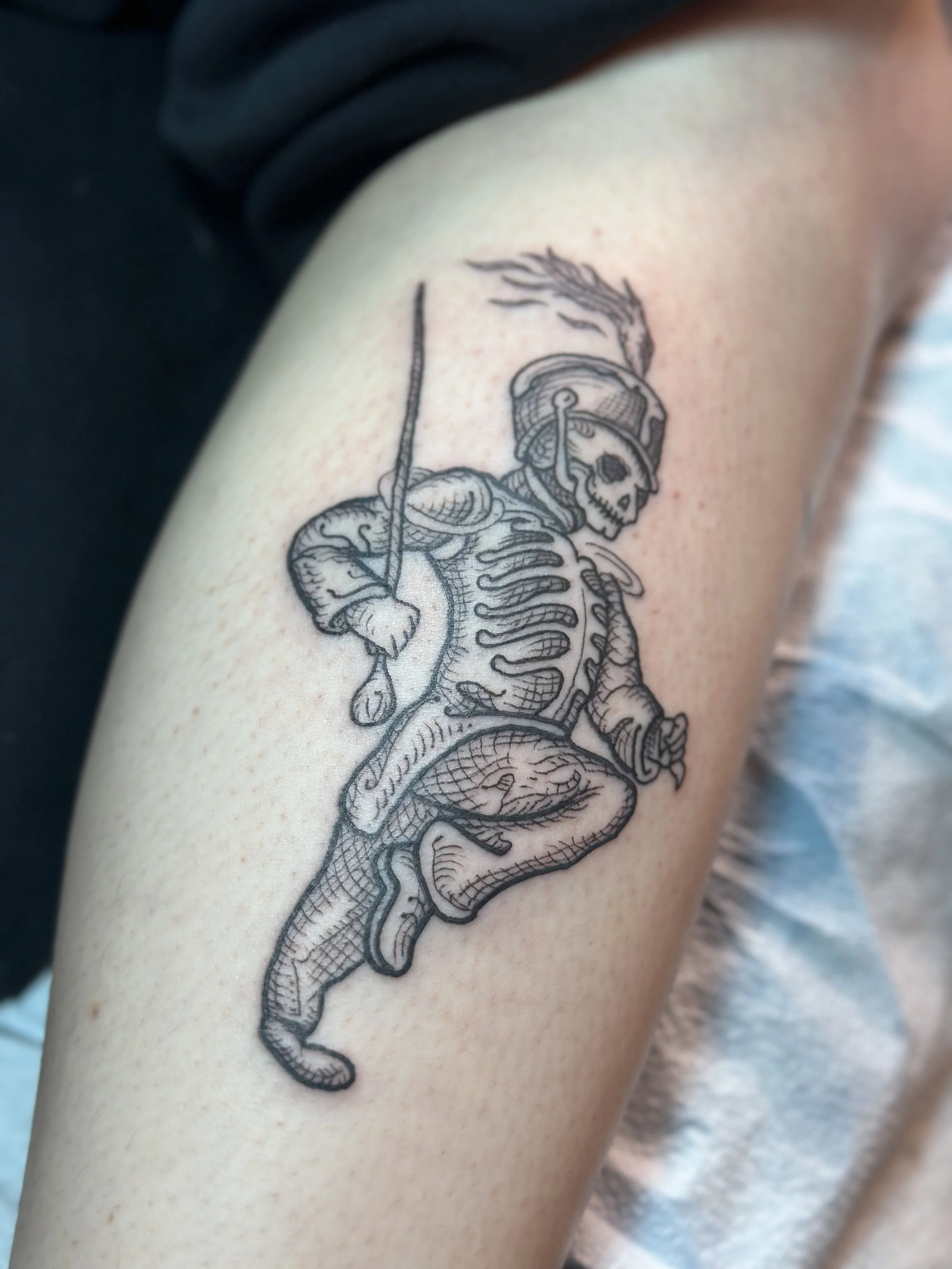 Black and gray tattoo of a skeleton soldier wearing a helmet with a feather, holding a spear, and riding a mythical creature on a person's arm.