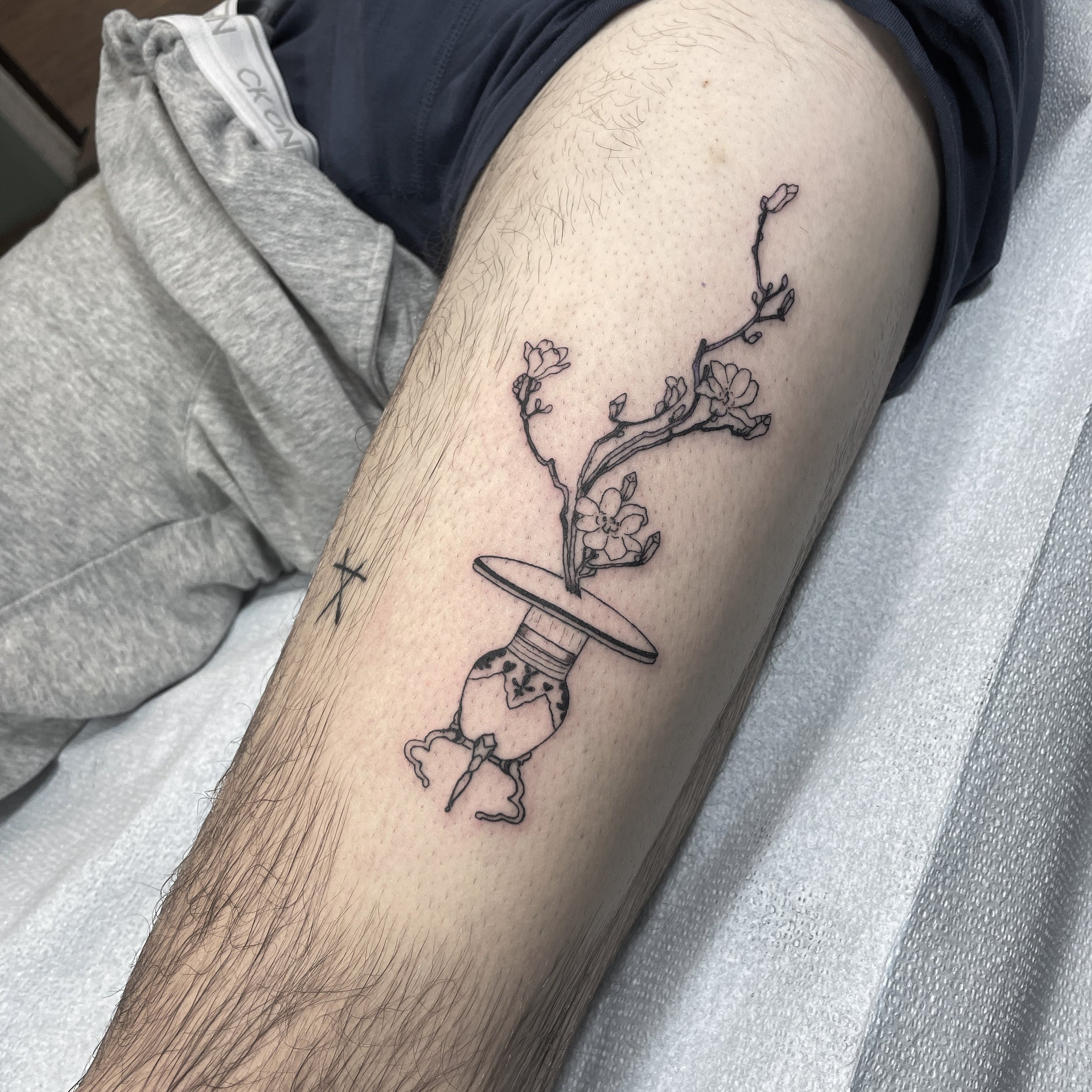 Line art tattoo of a teapot with a branch of blossoms emerging from its spout and lid, on a person's inner arm.