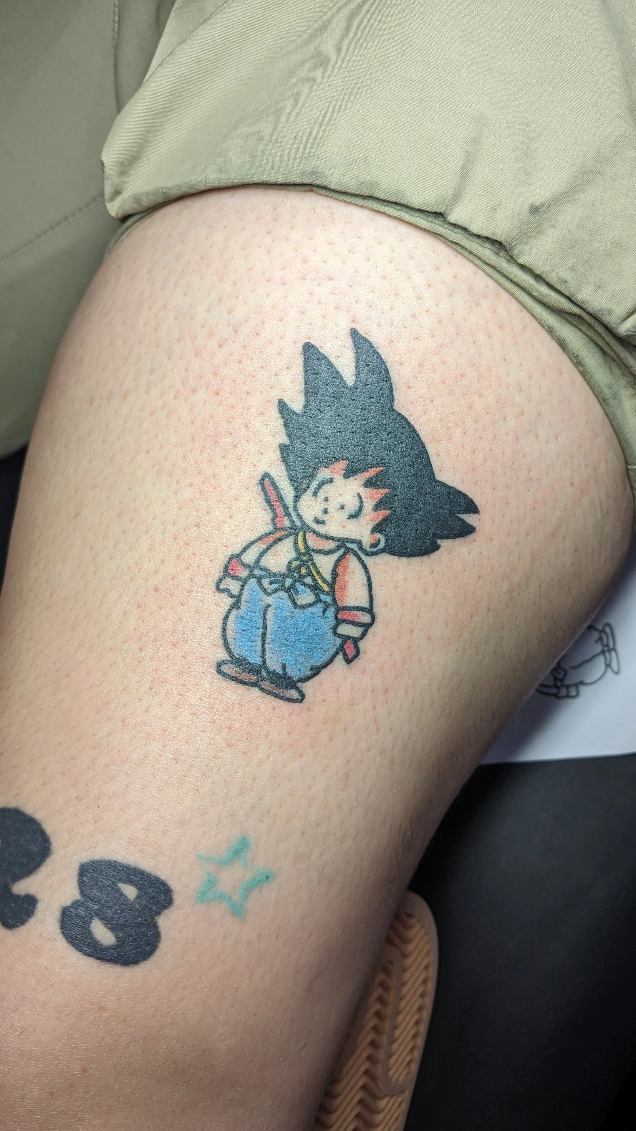 Tattoo of a chibi-style character with spiky black hair, wearing a red shirt, blue overalls, and carrying a sword on the back.
