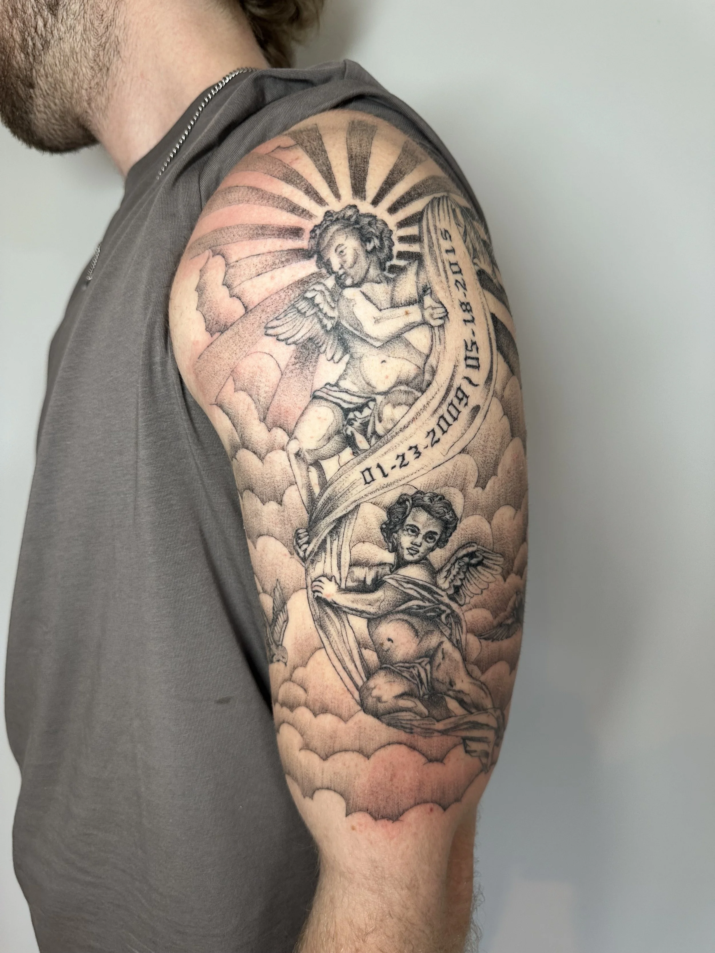 Tattoo on man's upper arm featuring two cherubs, clouds, and a banner with the date 01.23.18 to 02.28.2015.
