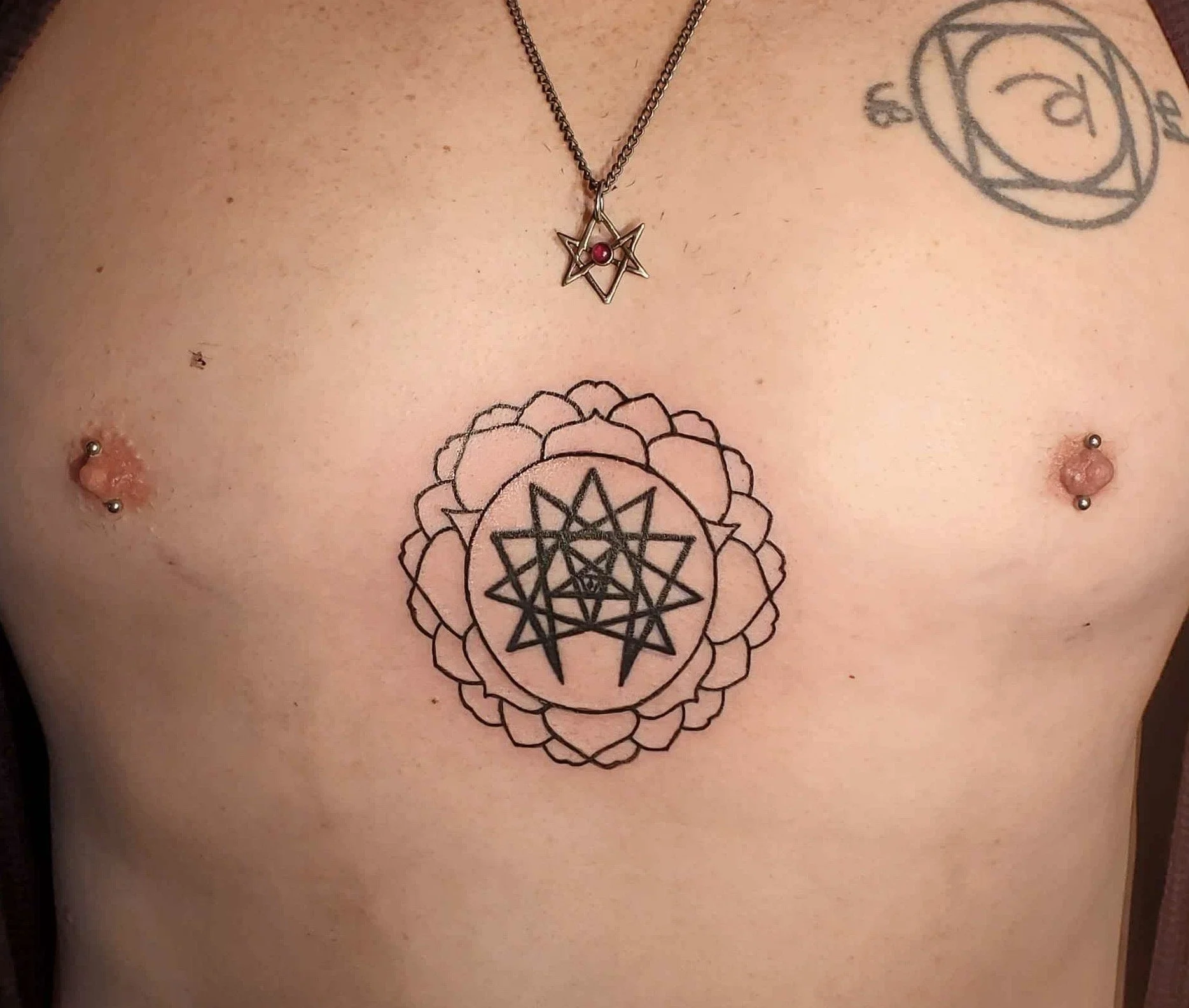 A person's bare chest with tattoo designs, piercings, and a necklace. The tattoos include a geometric star surrounded by a floral pattern on the stomach and a circular design on the shoulder. 