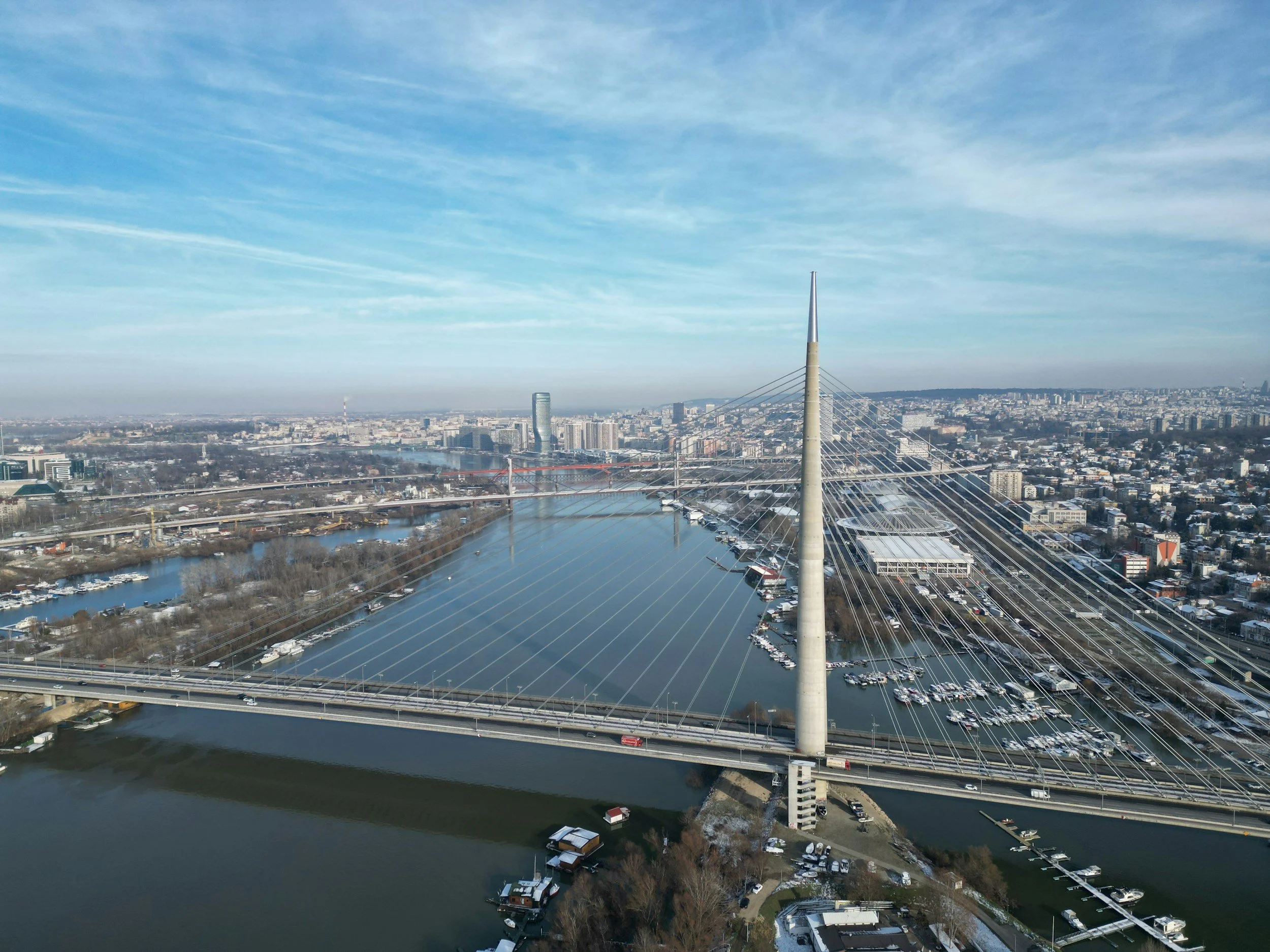 Aerial view of a city with a cable-stayed bridge crossing a wide river, showing boats and surrounding urban landscape under a blue sky with clouds.
