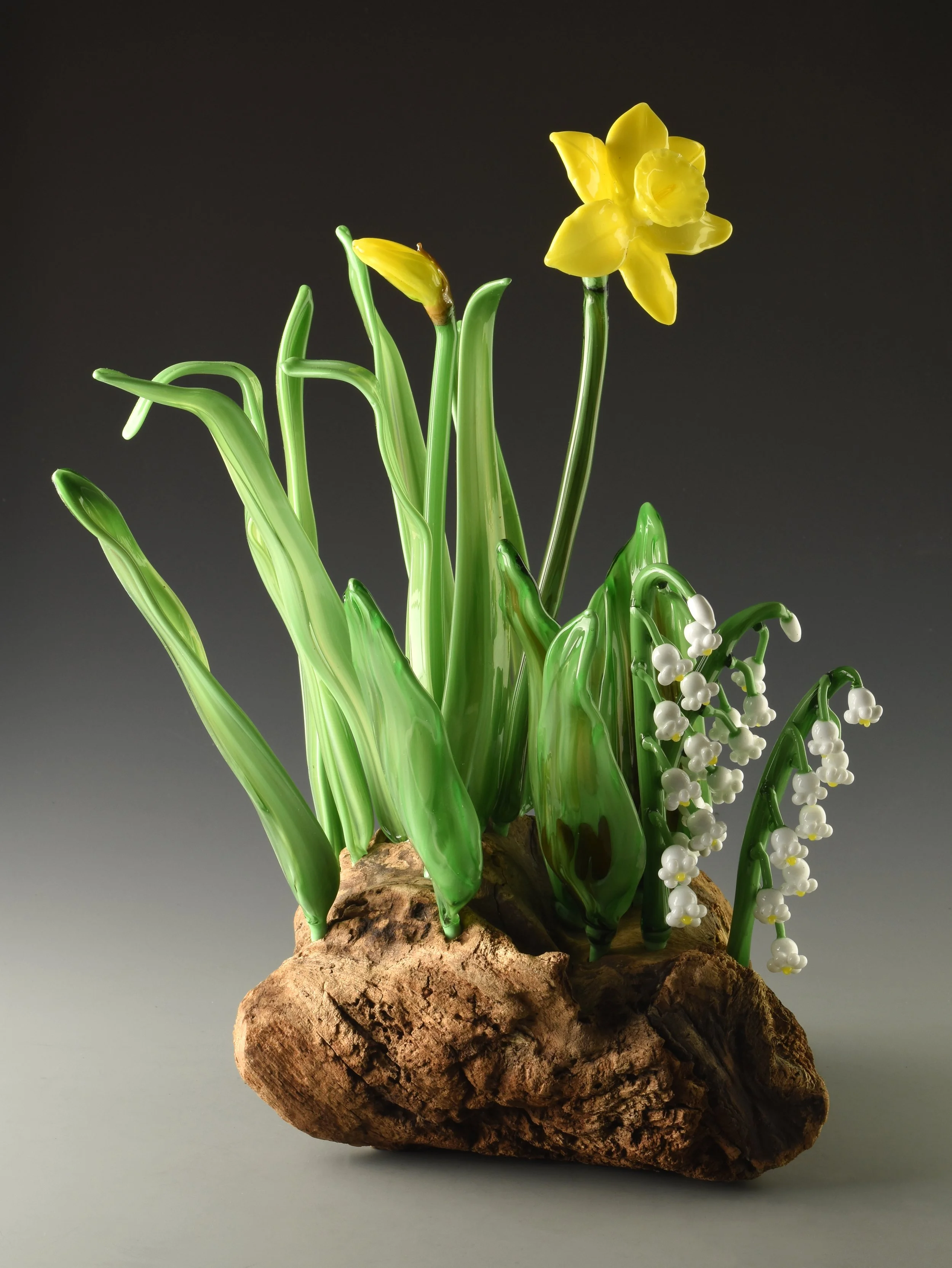 Artificial yellow daffodil and white lily of the valley flowers on a rocky base against a dark to light gray gradient background.