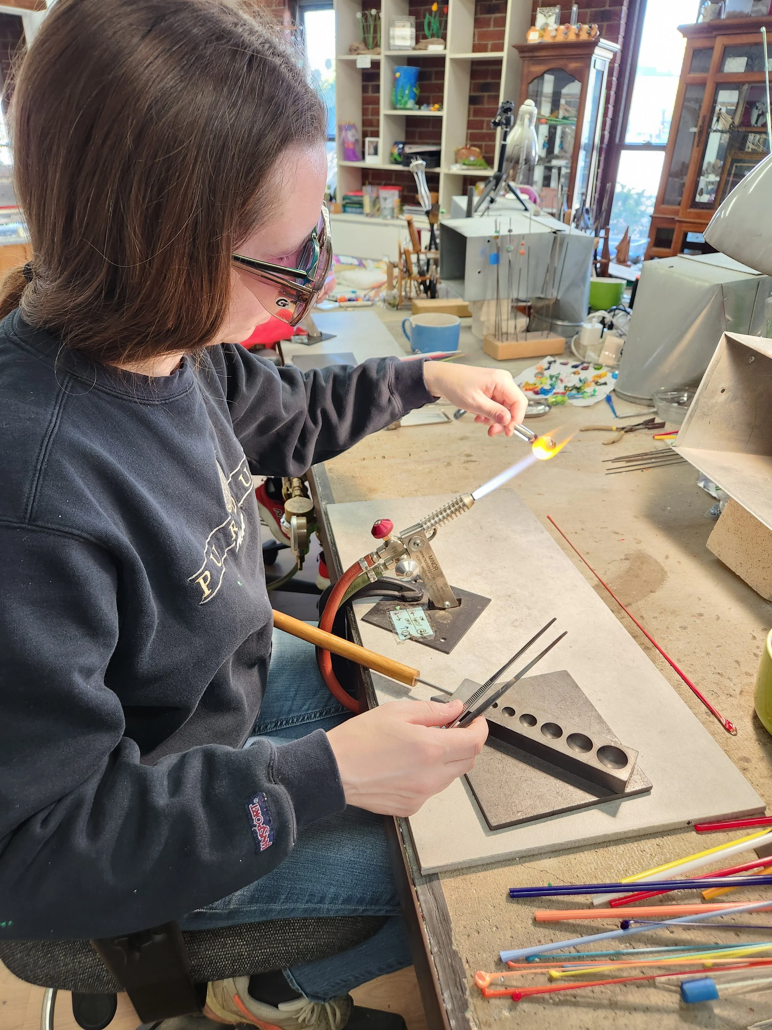 A person working with a torch and tools at a workbench, surrounded by glass rods and equipment in a workshop, possibly for glassblowing or jewelry making.