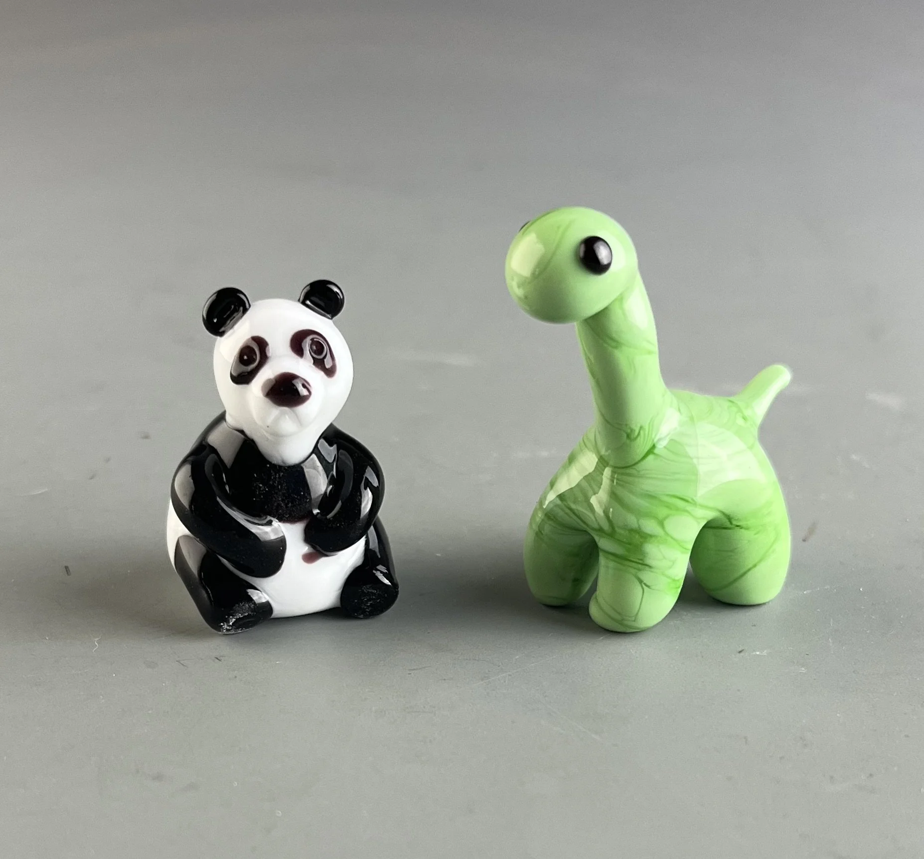 Photo of two glass animal figurines, a panda and a dinosaur, placed on a gray surface against a plain gray background.