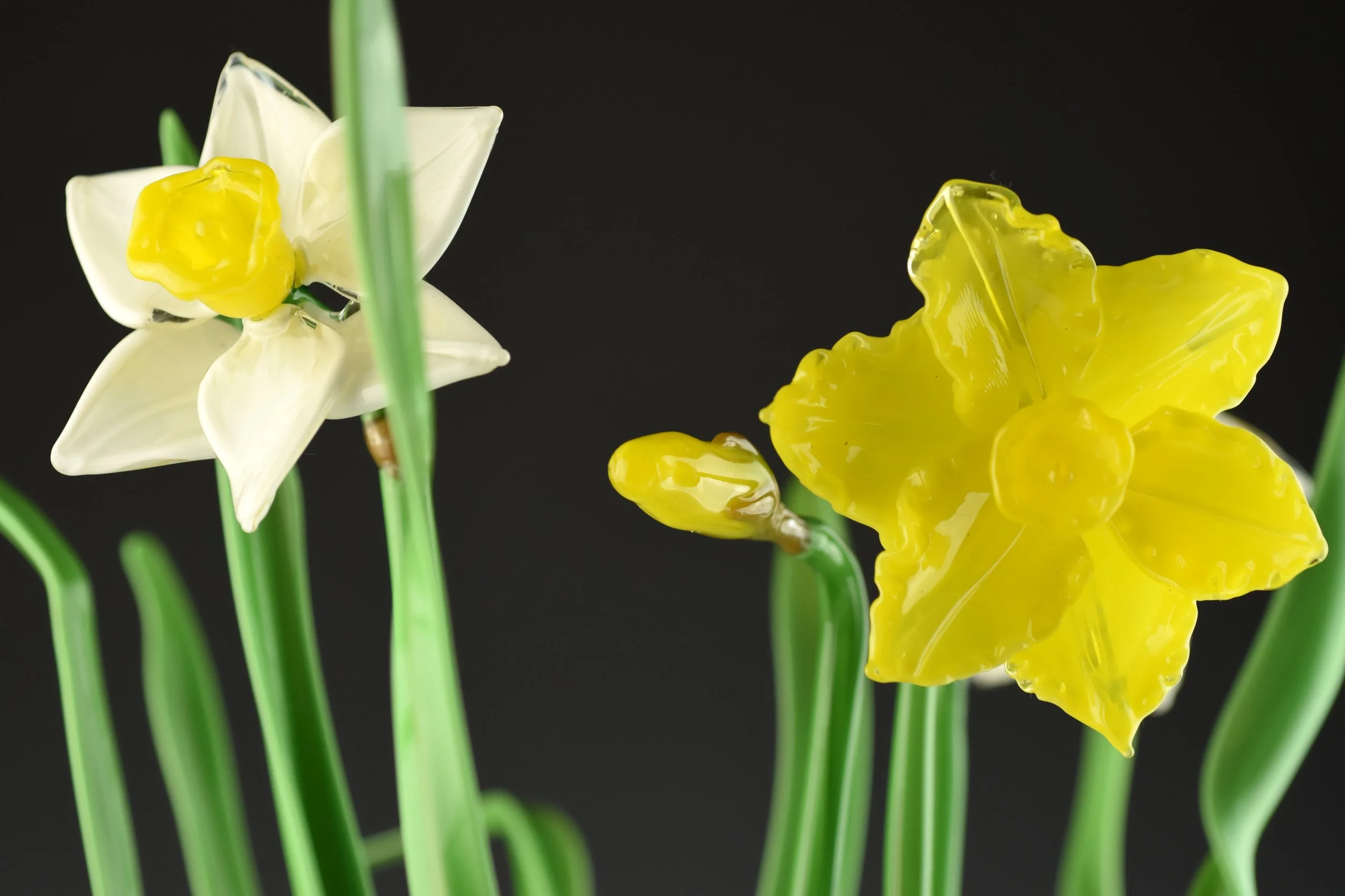 Close-up of colorful glass flowers, including white and yellow daffodils, with green stems, on a dark background.