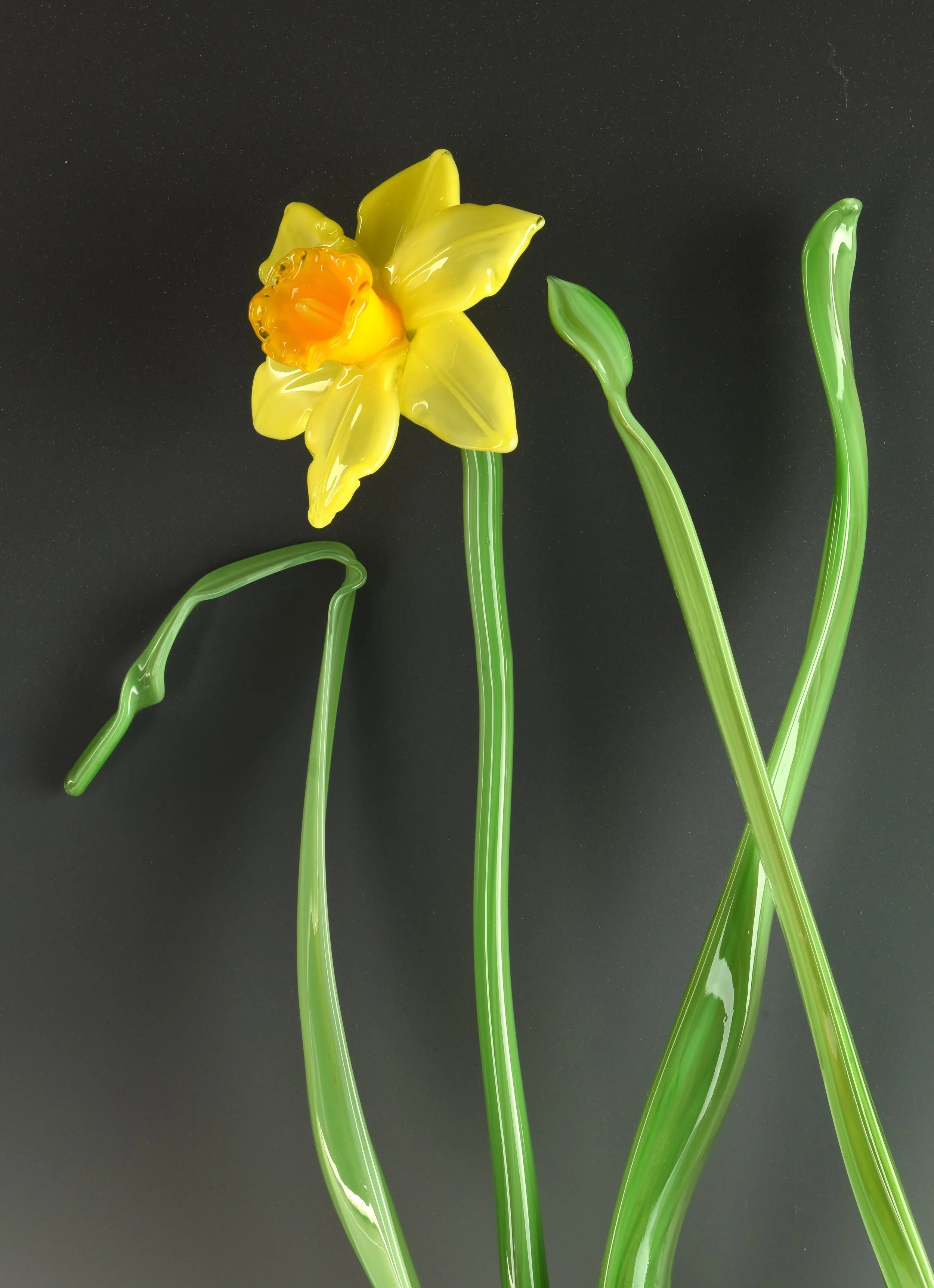 A glass sculpture of a yellow daffodil flower with a bright orange center, green stem, and leaves, set against a dark background.