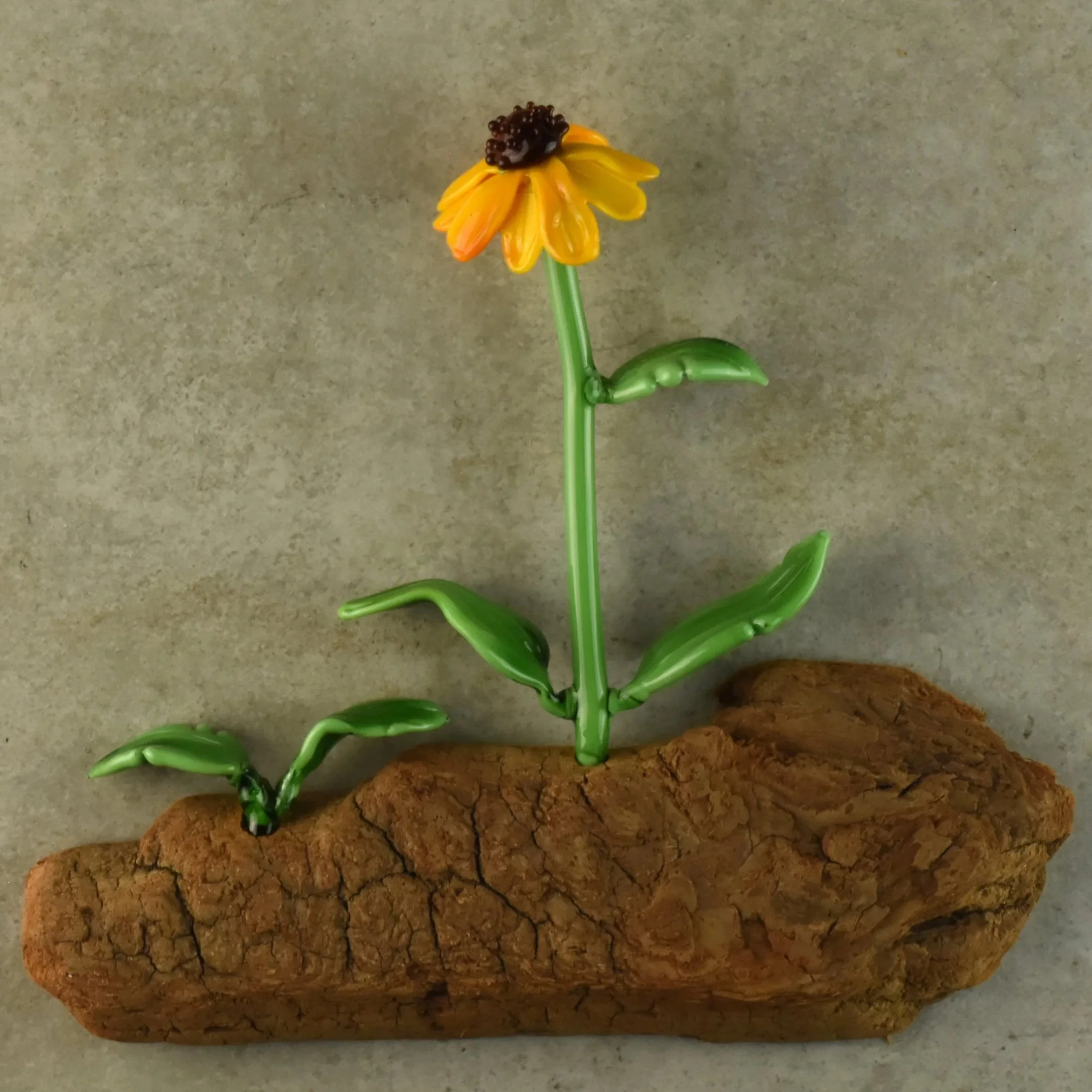 A yellow artificial flower with a dark brown center and green leaves, mounted on a green stem, resting on a piece of textured bark, against a plain gray background.